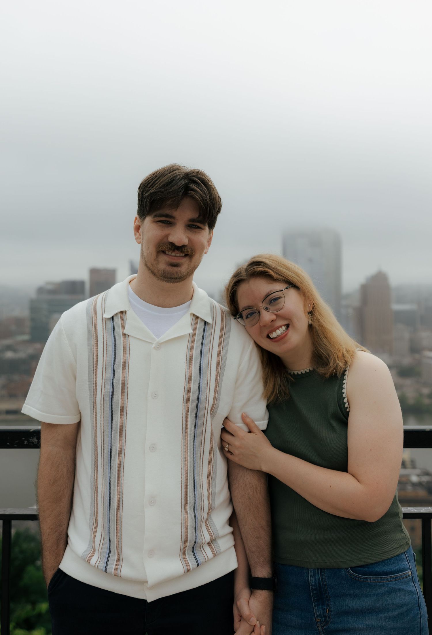 A smiling couple poses together on a rooftop with a city skyline in the background on a cloudy day.