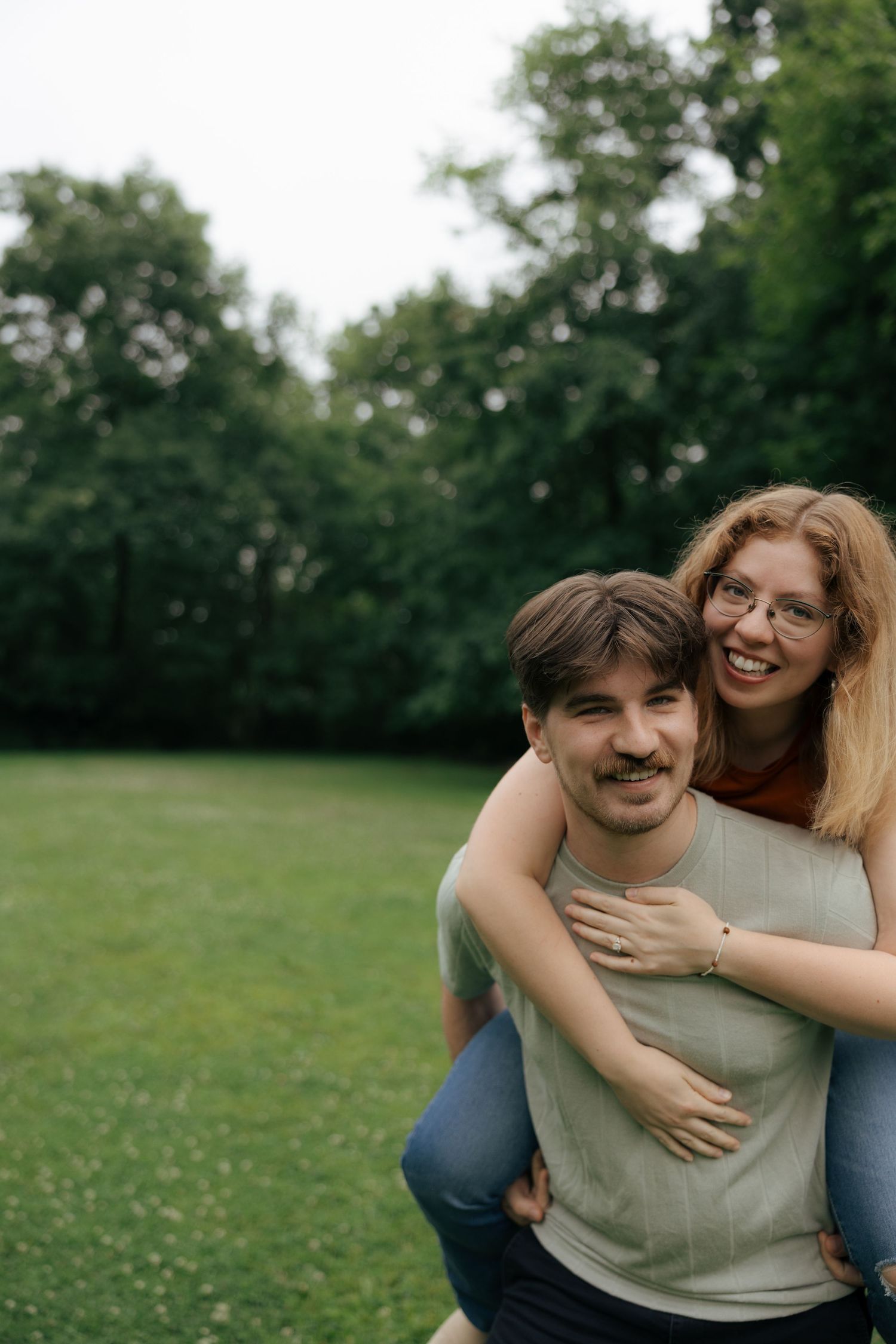 A smiling woman with glasses rides piggyback on her partner in a lush green park setting.