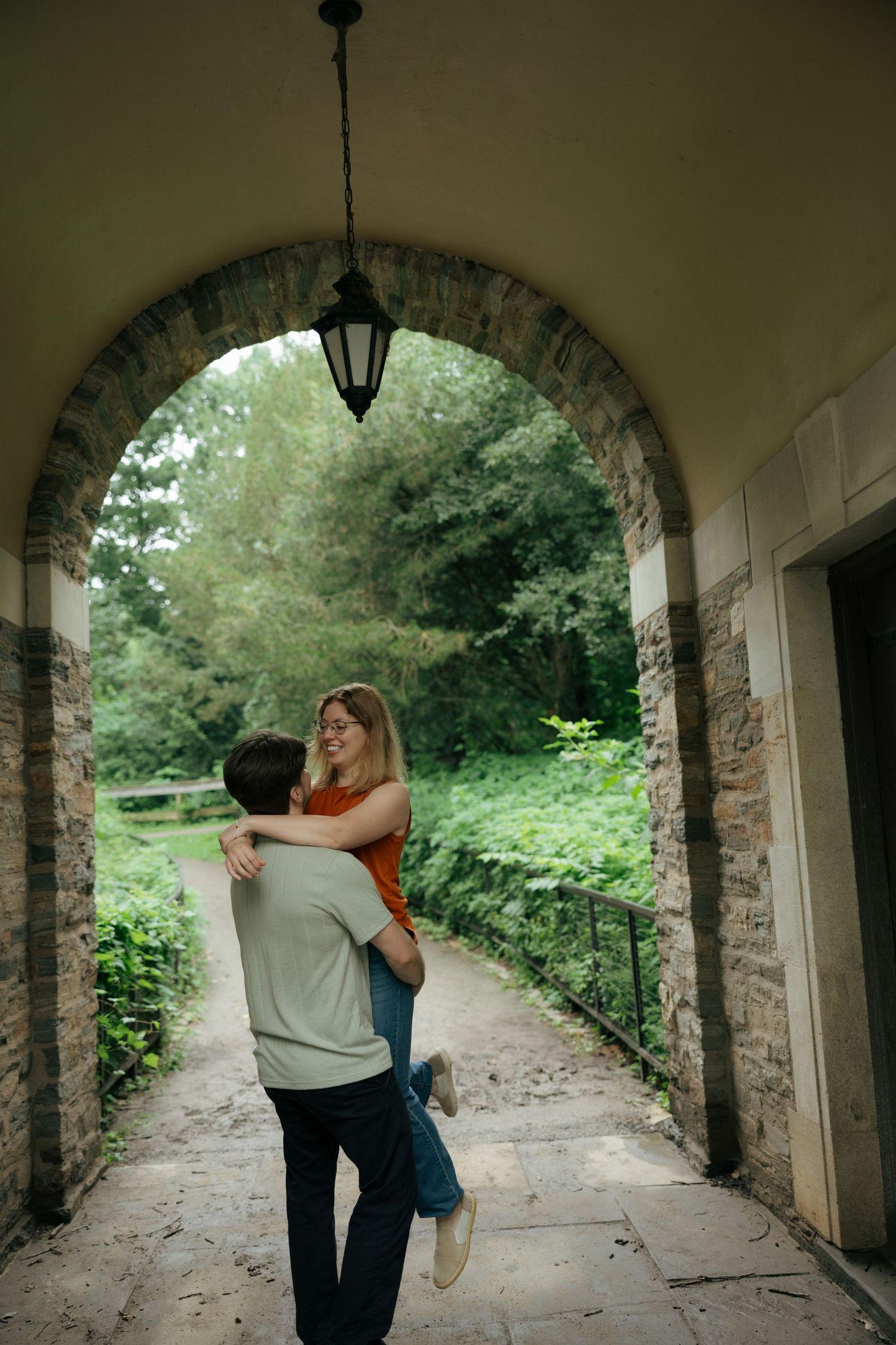 Couple sharing a joyful embrace under a stone archway surrounded by lush green trees in a park.