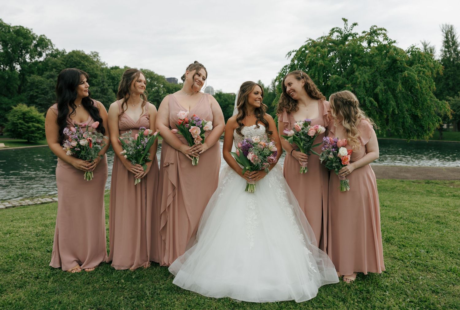 Bride in white gown poses with five bridesmaids in dusty rose dresses holding floral bouquets outdoors.