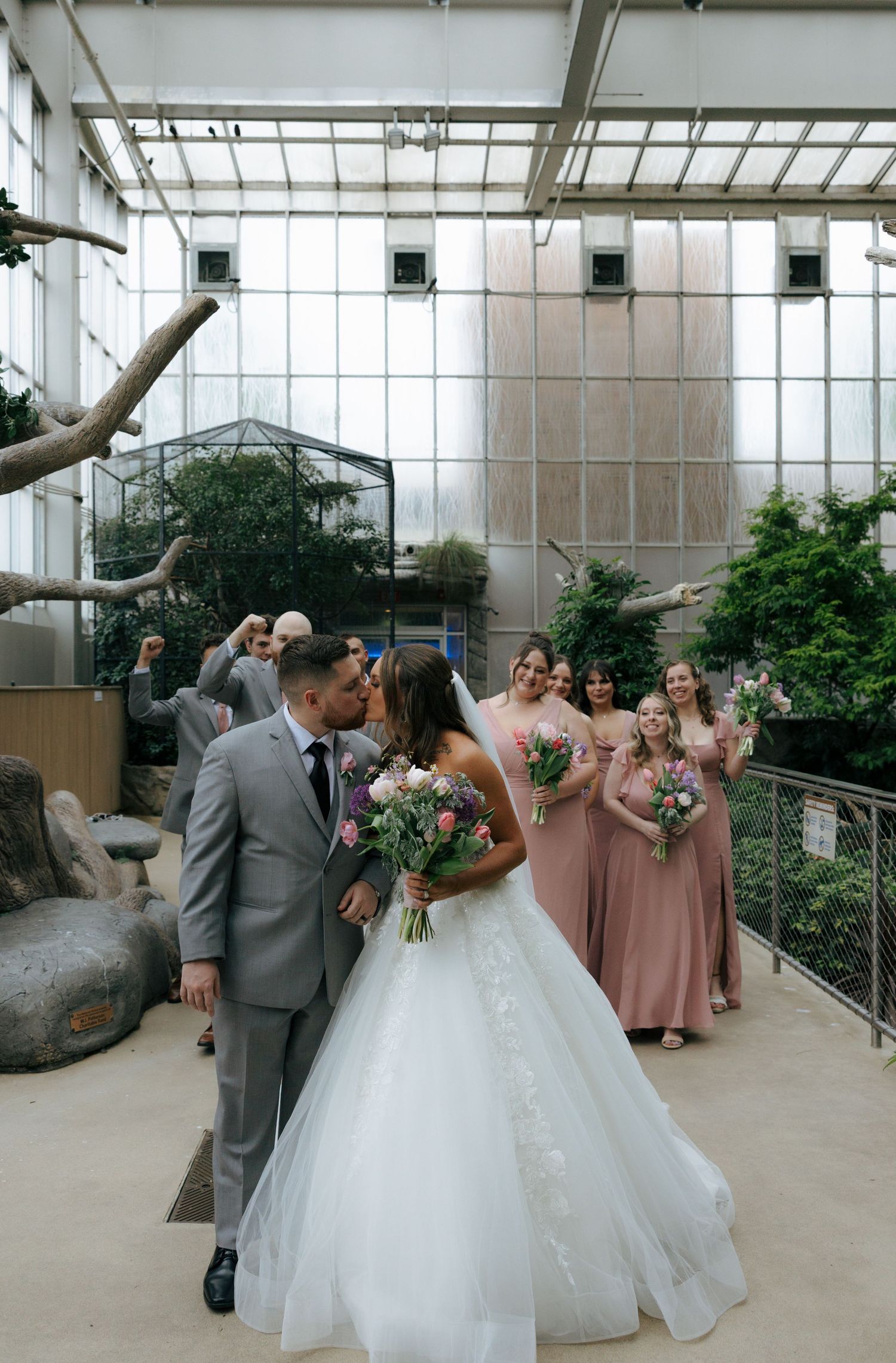 Bride and groom share a kiss while bridesmaids in dusty rose gowns celebrate inside a sunlit botanical garden venue.