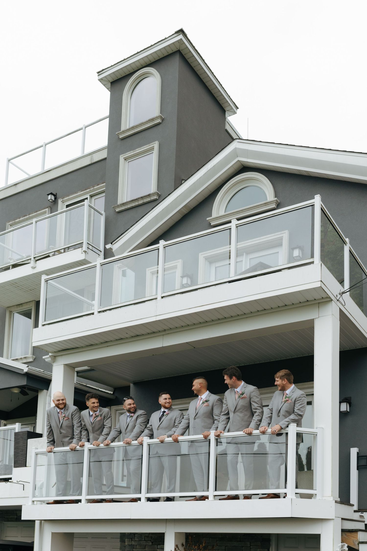 Groomsmen in gray suits stand together on a white balcony of a modern gray building during a wedding celebration.