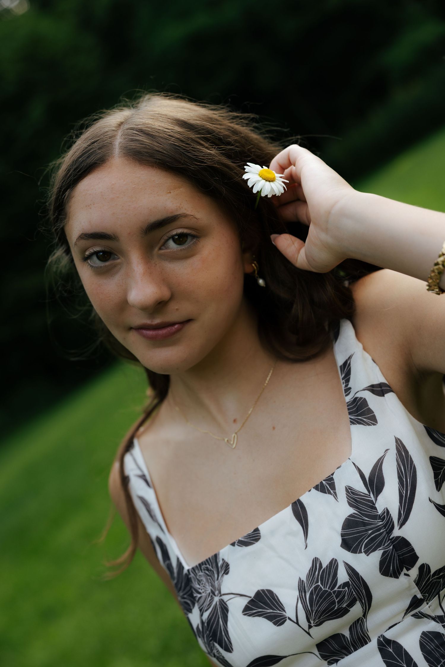 Young woman with daisy in hair wearing black and white floral dress, posing in green garden.