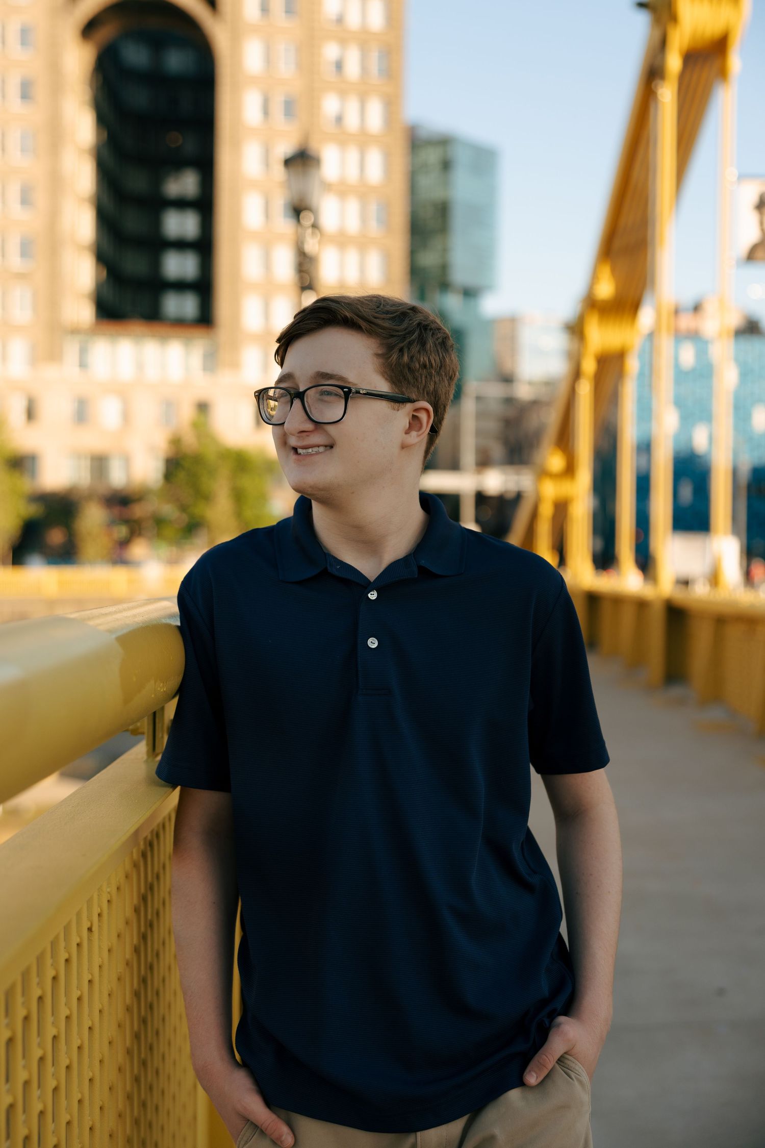 Teen boy in navy polo and glasses smiling on a yellow bridge with city buildings in background.