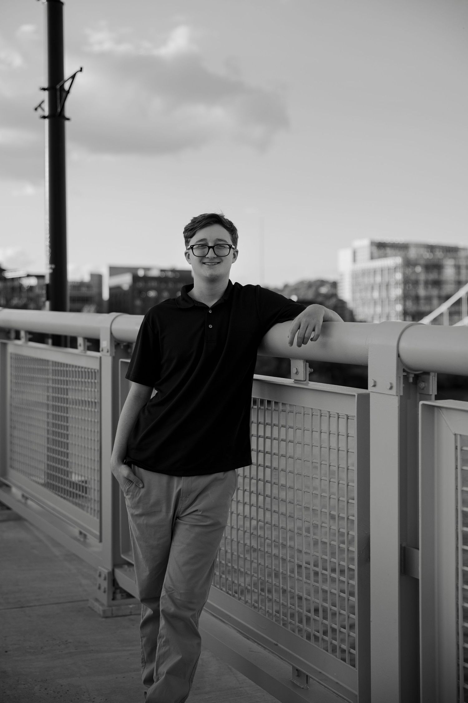 Young man in black polo and glasses leaning on bridge railing, black and white urban portrait photo.