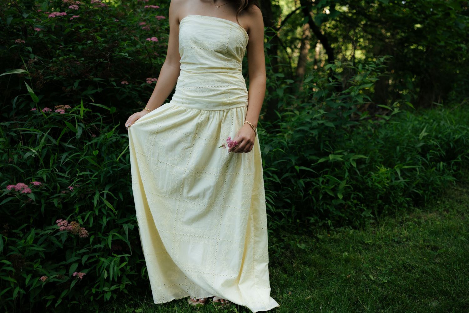 Woman in long yellow strapless gown holding pink flower in garden.