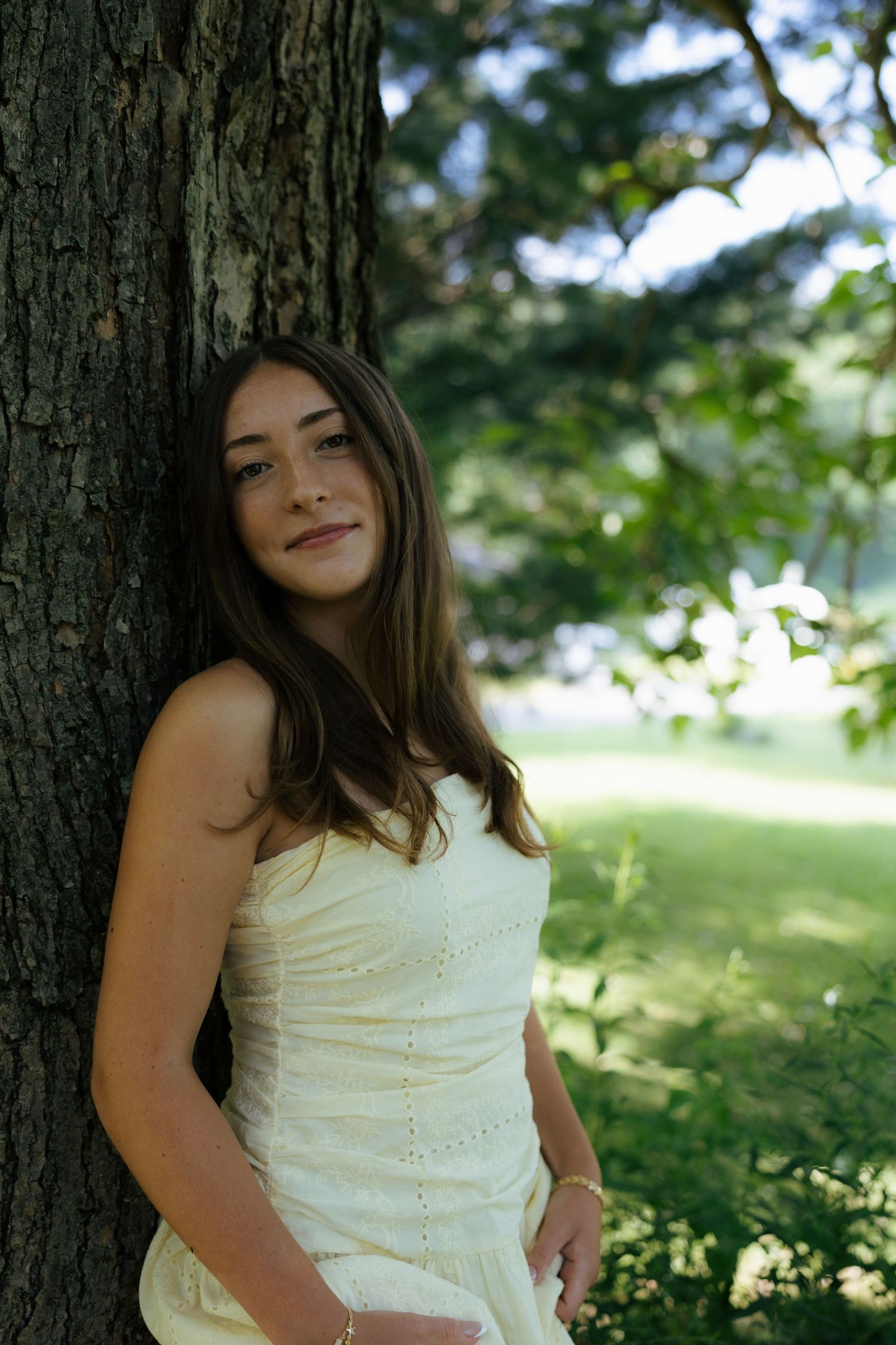 Smiling young woman in a strapless cream dress posing against a large tree in a green outdoor setting.