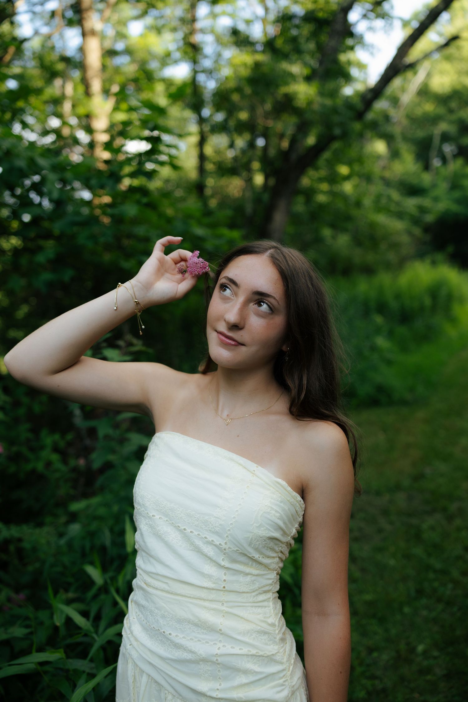 Elegant young woman in cream strapless dress holding pink flower in green forest setting.