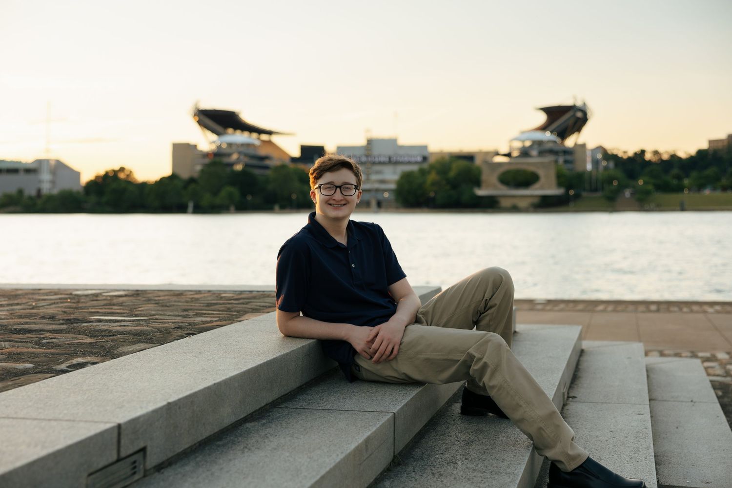 Teen boy with glasses in navy polo sits on steps by waterfront at dusk near stadium.