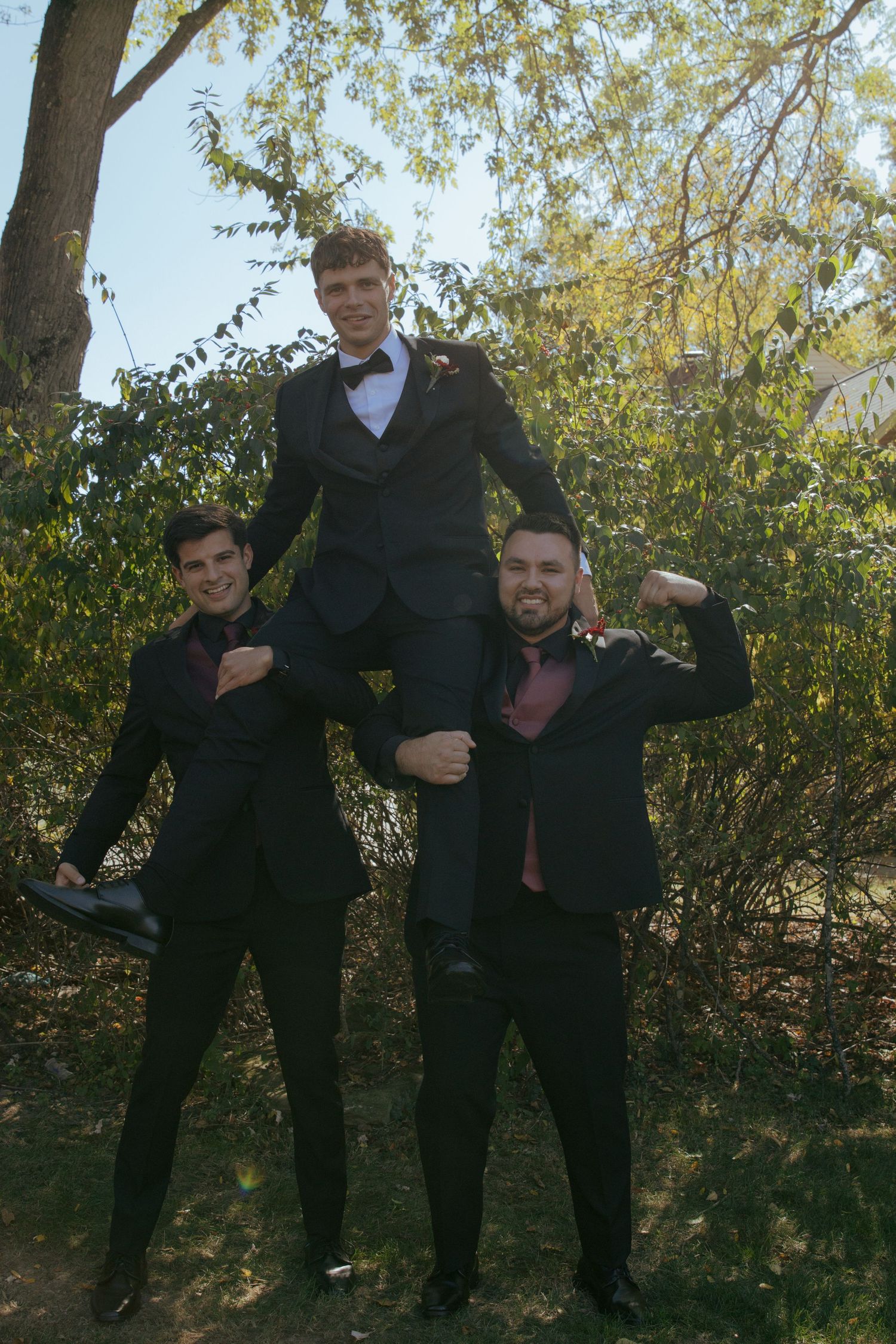 Three young men in black tuxedos pose outdoors, with one man lifted on the shoulders of the other two.