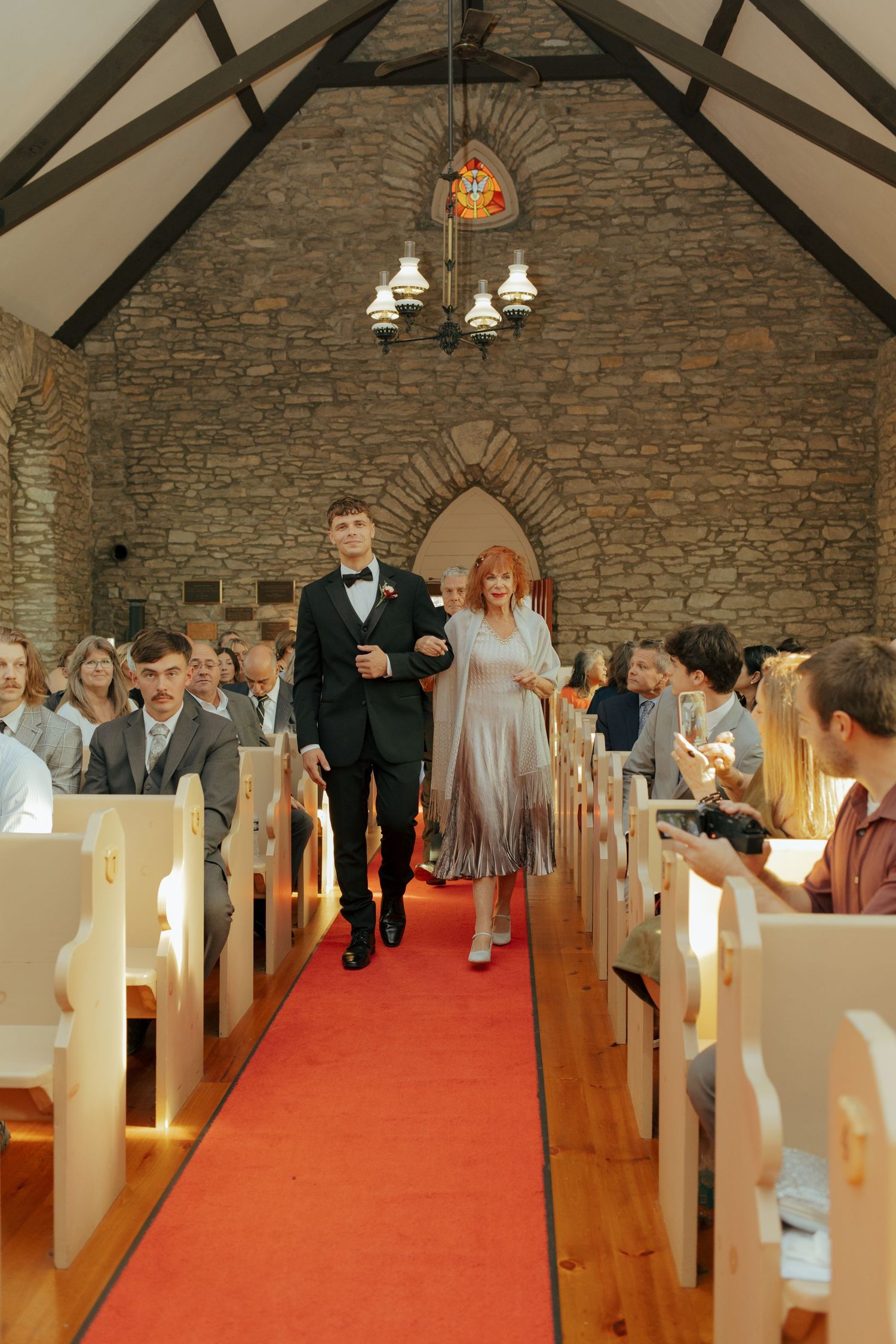 Groom in tuxedo walks down red carpet aisle with elegantly dressed woman in stone chapel wedding ceremony.