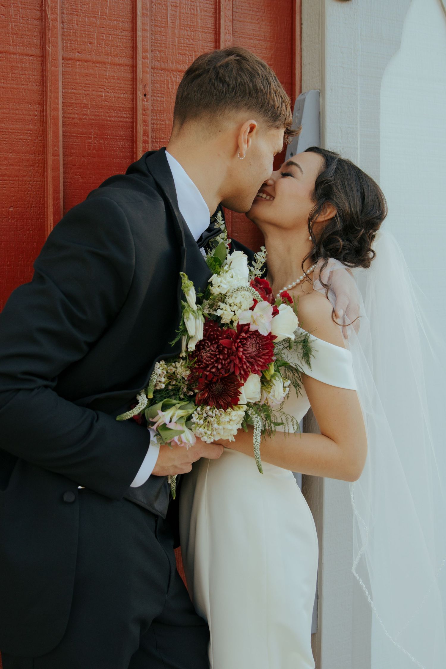 Bride and groom share a romantic kiss against a red wall, bride holding a stunning red and white bouquet.