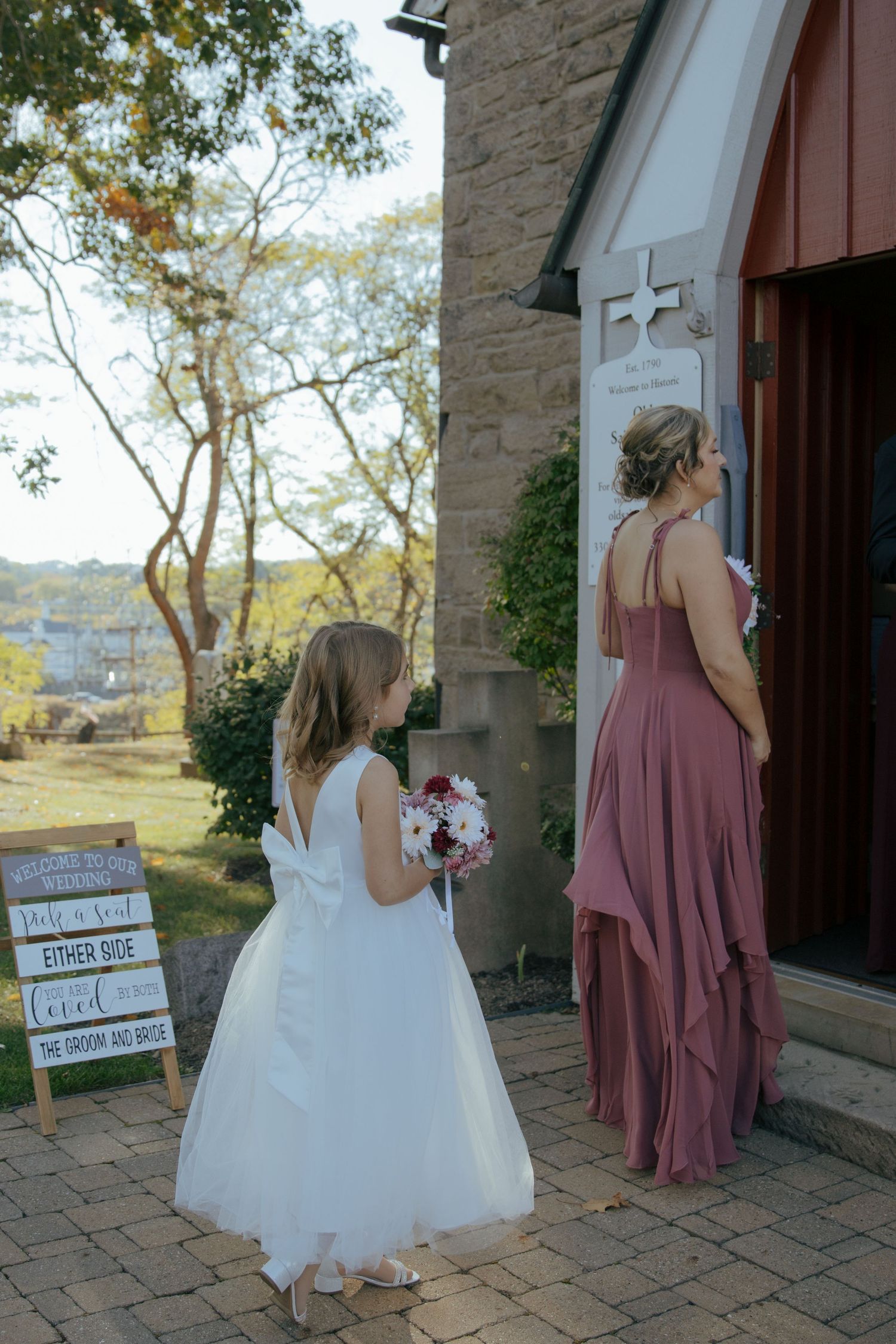 Flower girl in white dress and bridesmaid in dusty rose gown enter a charming stone church for wedding ceremony.