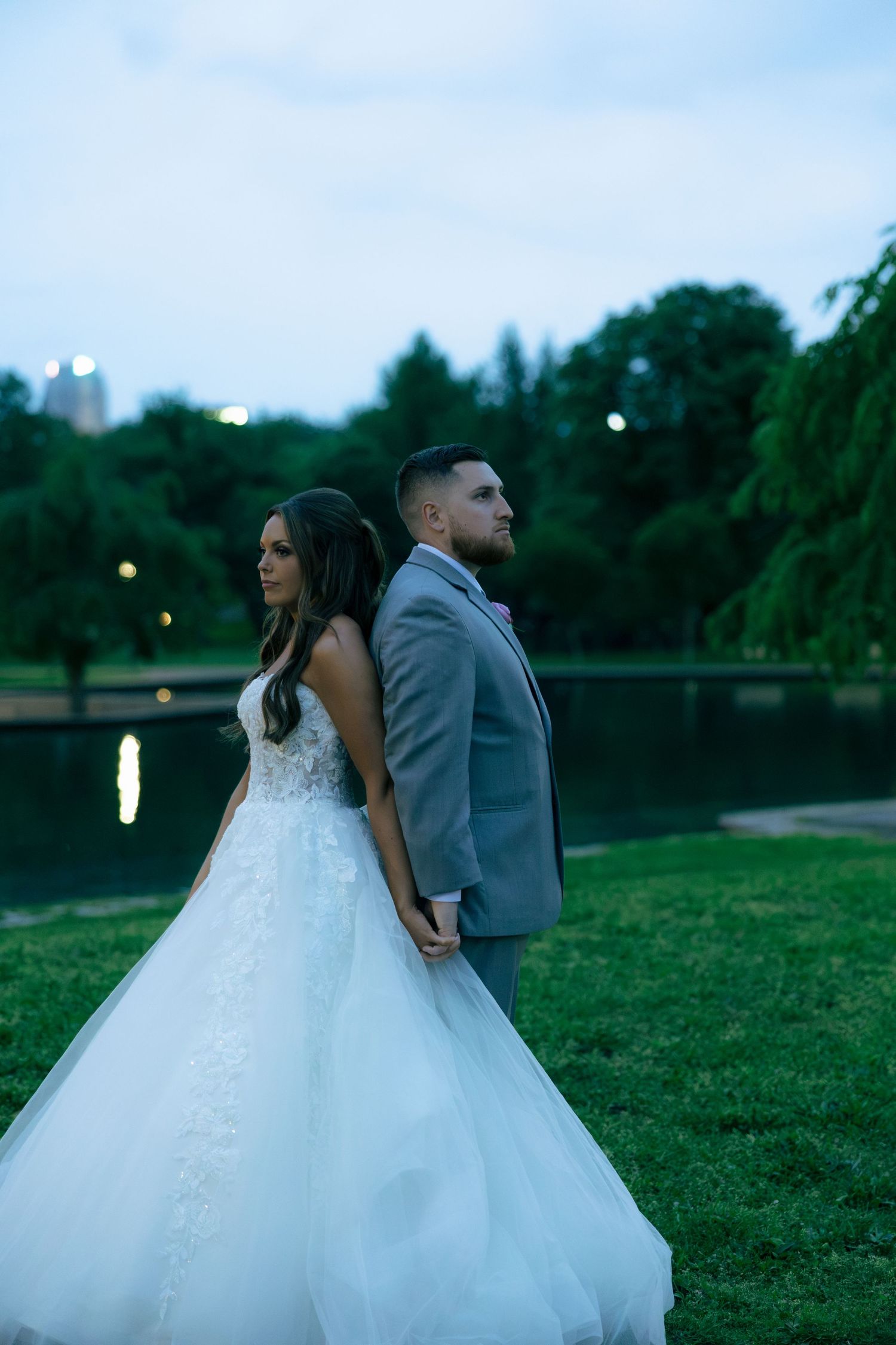 Bride and groom stand back-to-back holding hands by a serene park lake at dusk.
