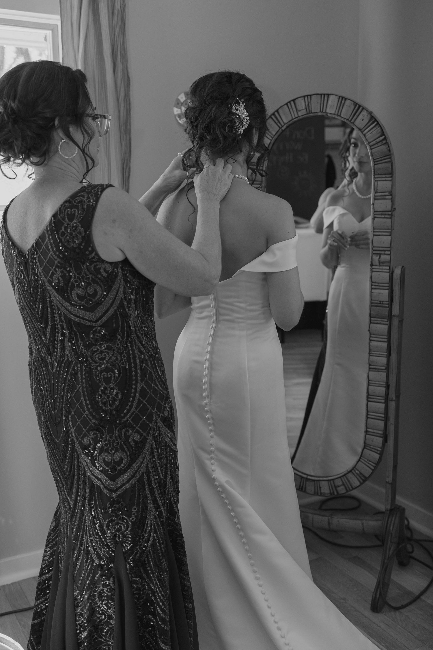 Bridesmaid in beaded gown helping bride in off-shoulder wedding dress fasten buttons before mirror.