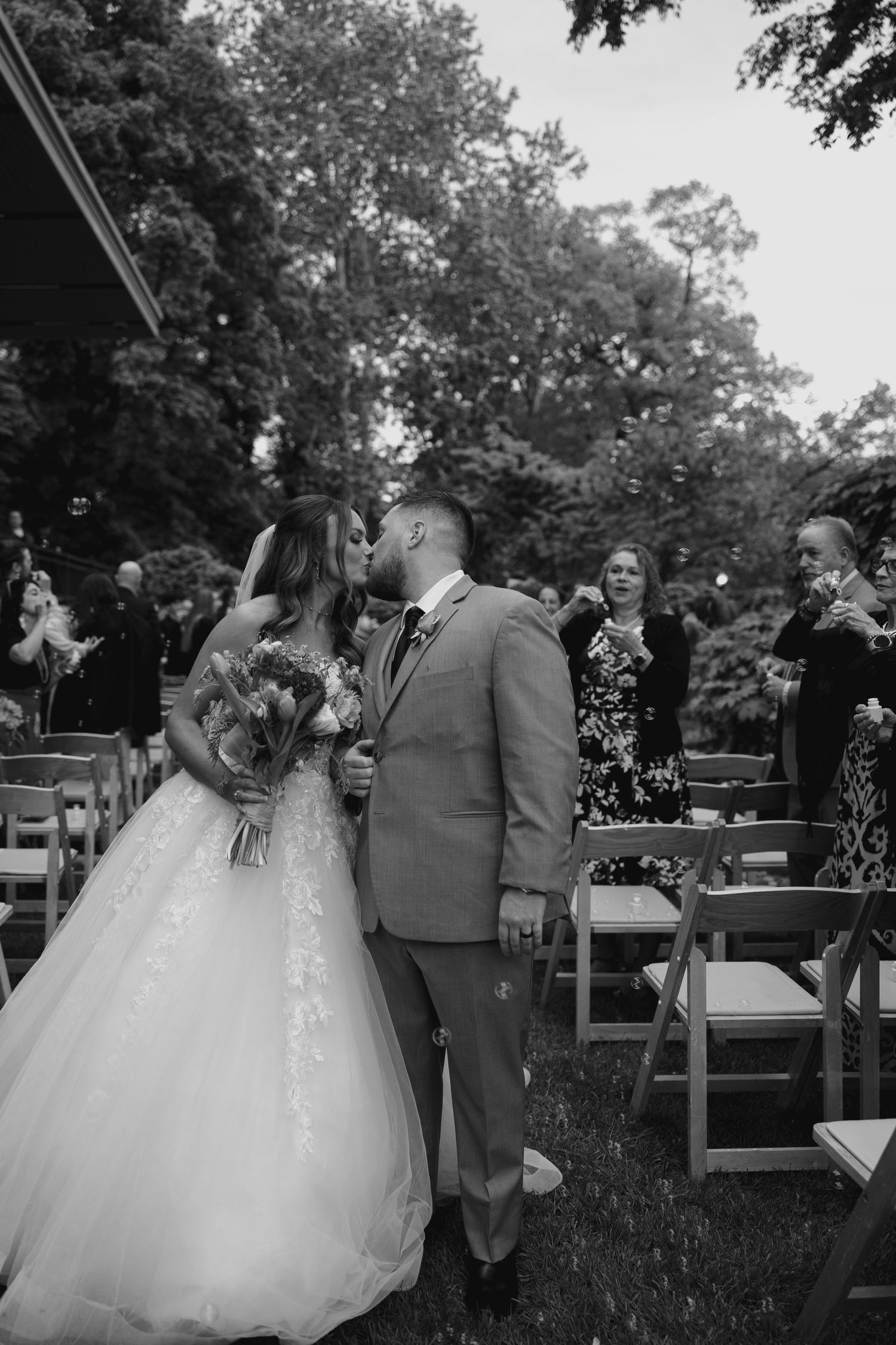 Black and white photo of newlyweds sharing first kiss at outdoor wedding ceremony after exchanging vows.