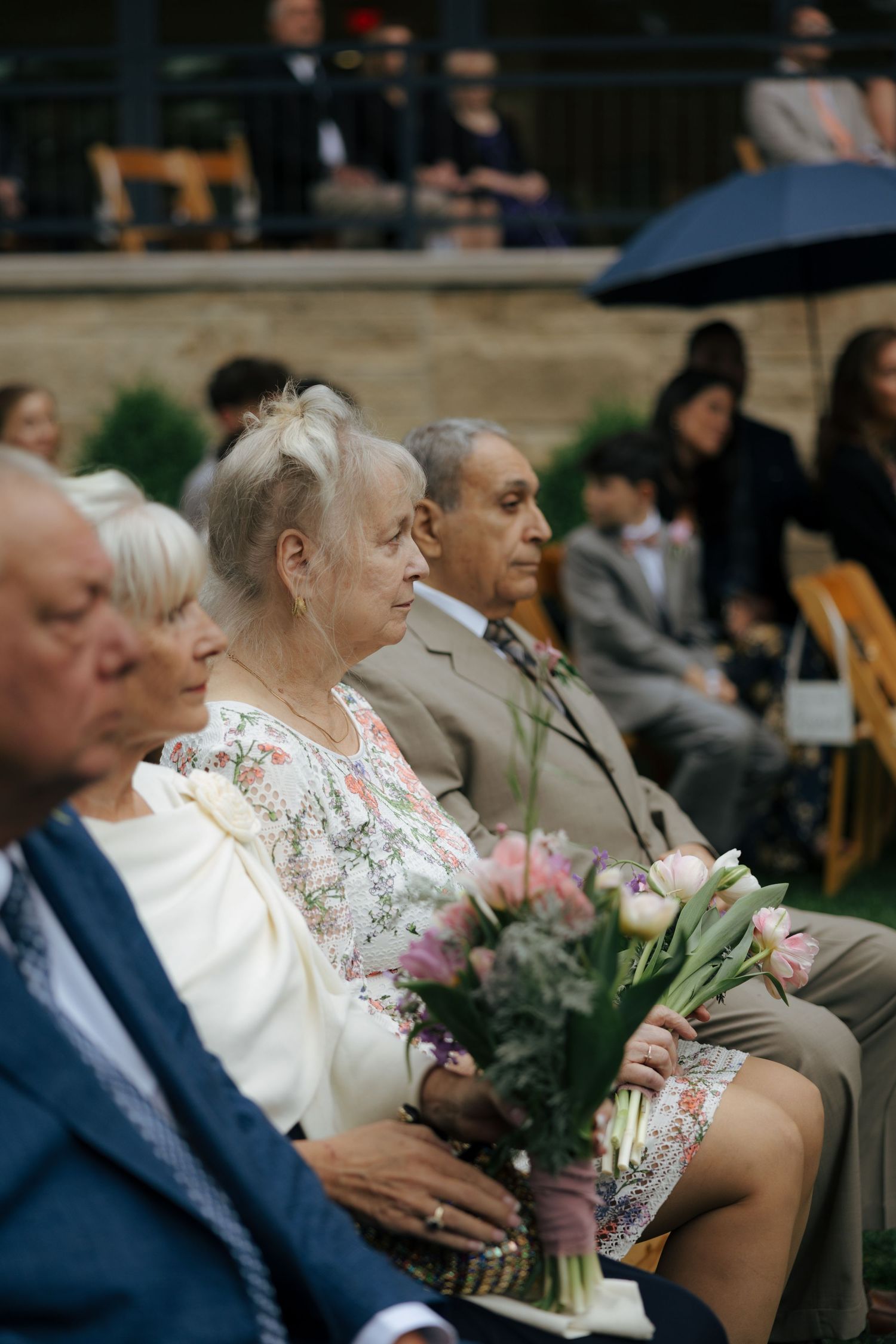 Wedding guests seated outdoors, elderly woman holding pink tulip bouquet during garden ceremony.