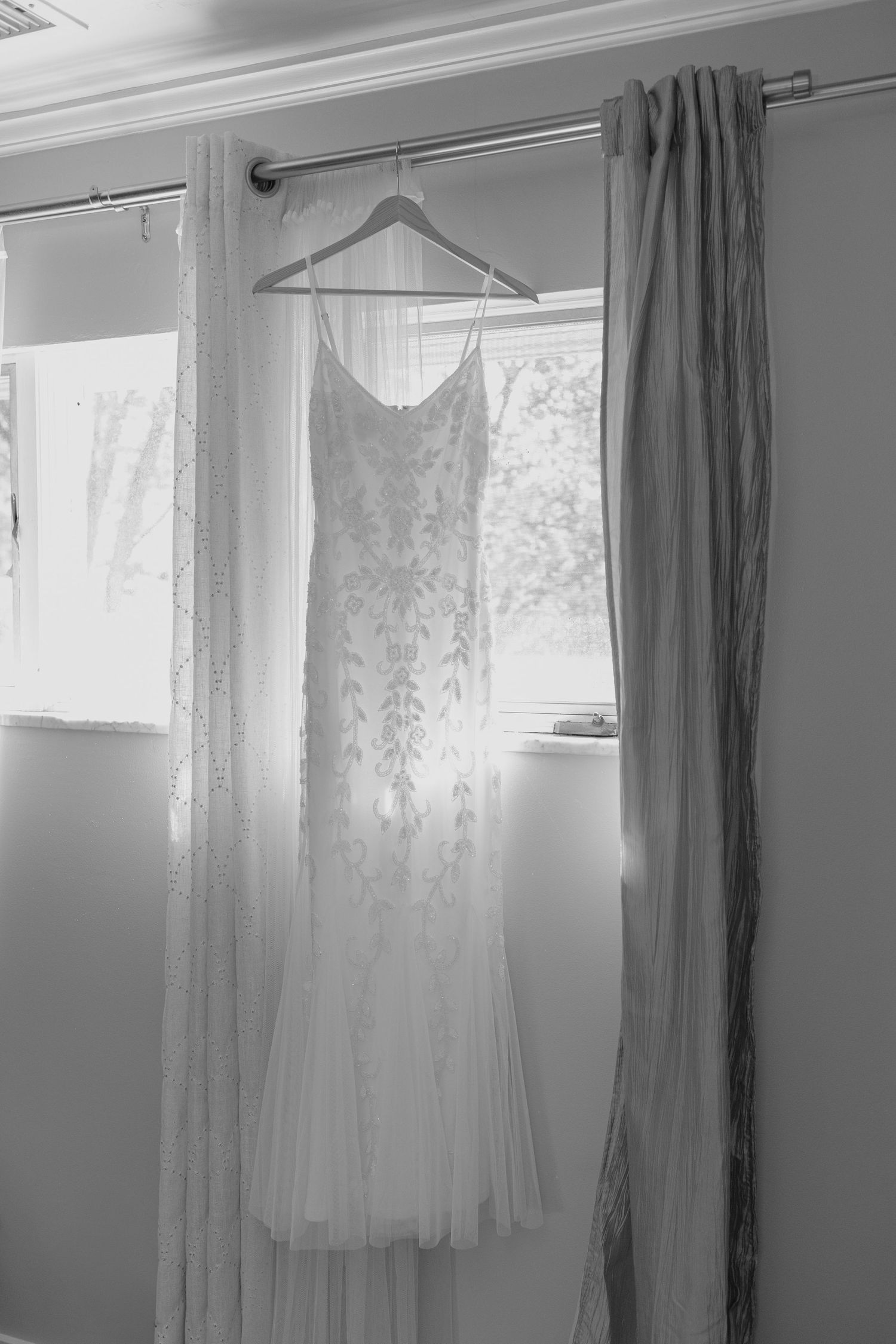 Elegant lace wedding dress hanging on a curtain rod in front of a bright window in black and white.