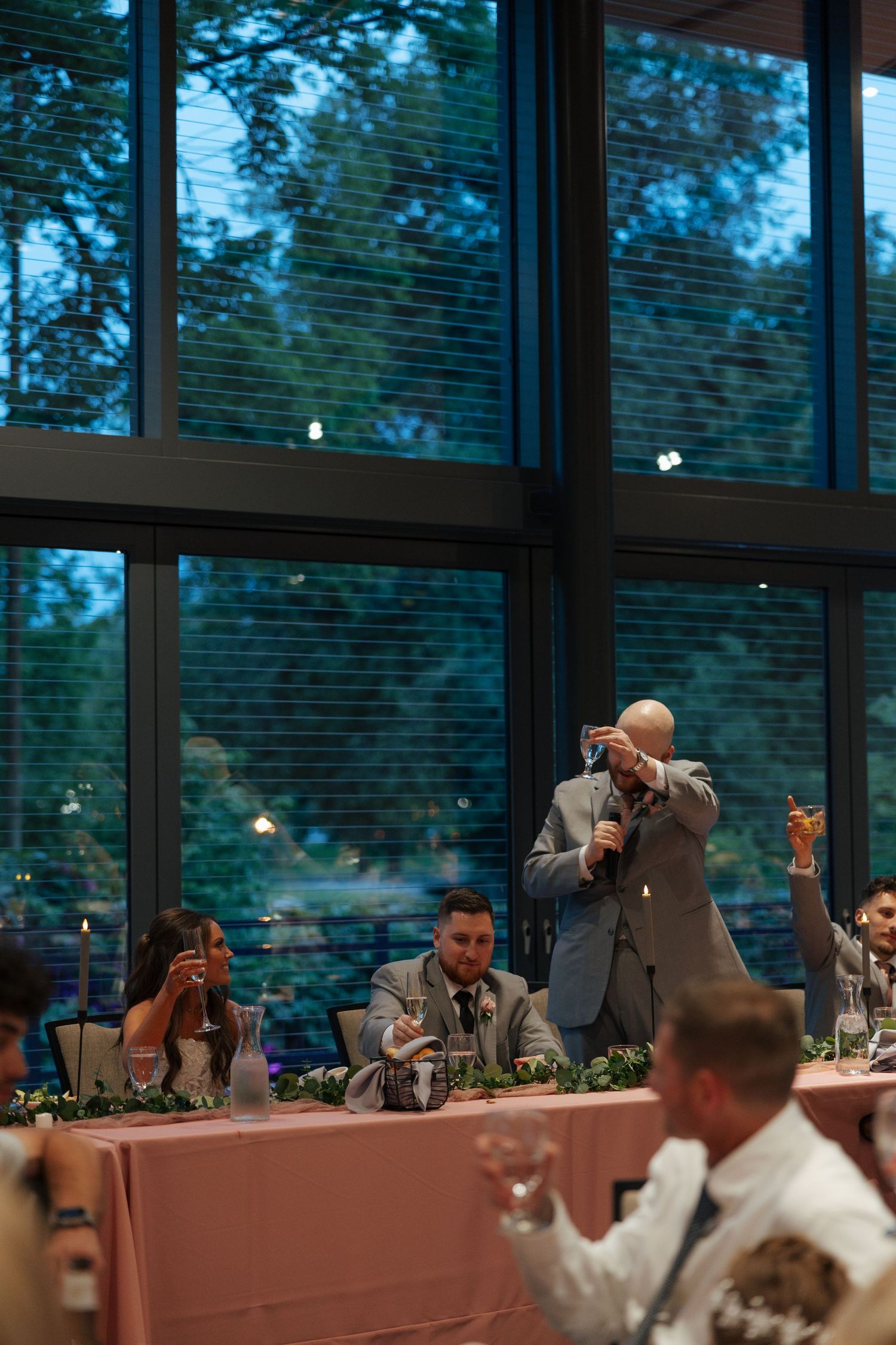 A man gives a toast at a wedding reception, raising a glass near large windows overlooking lush greenery.
