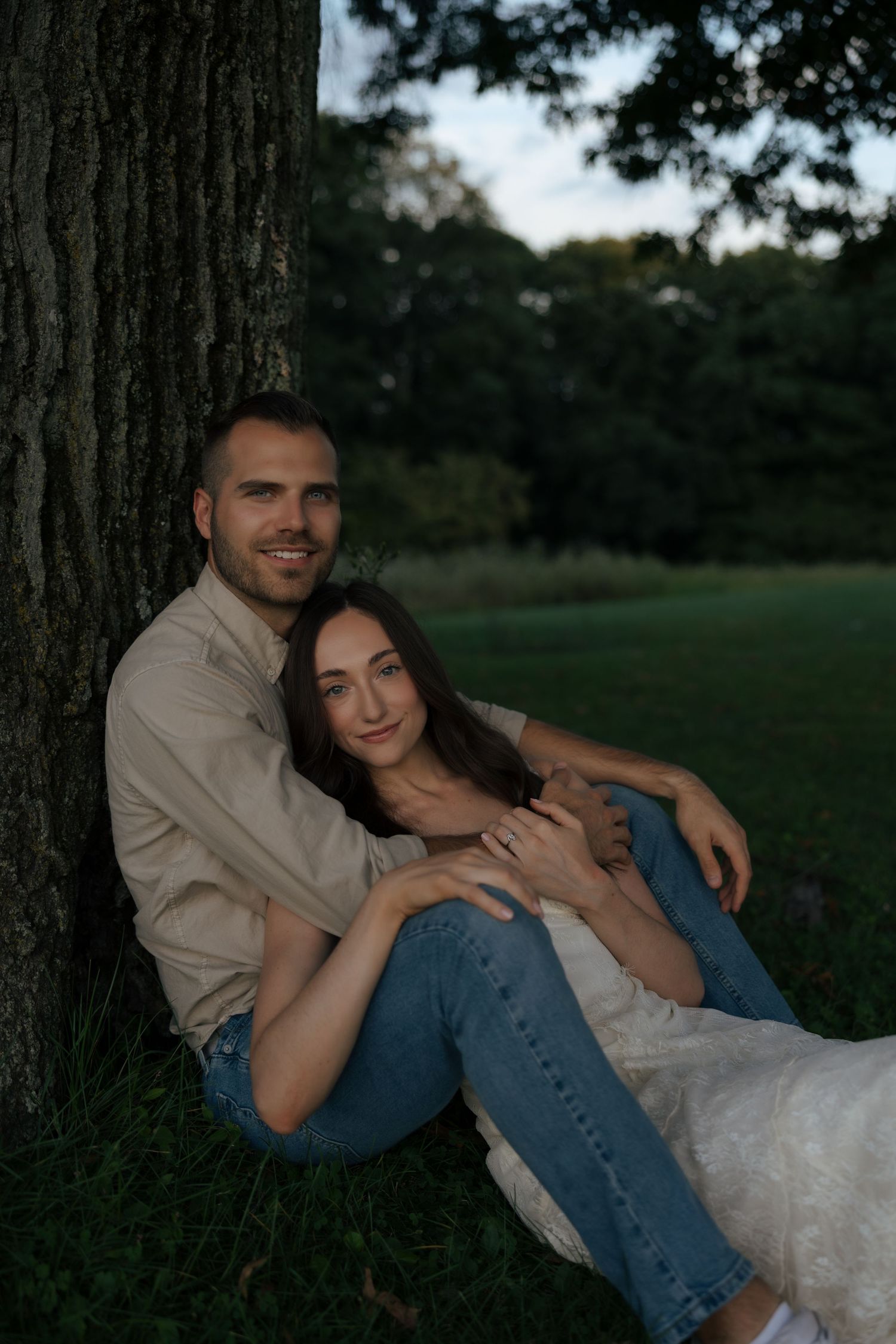 A happy couple sits together leaning against a tree trunk in a green park at golden hour.