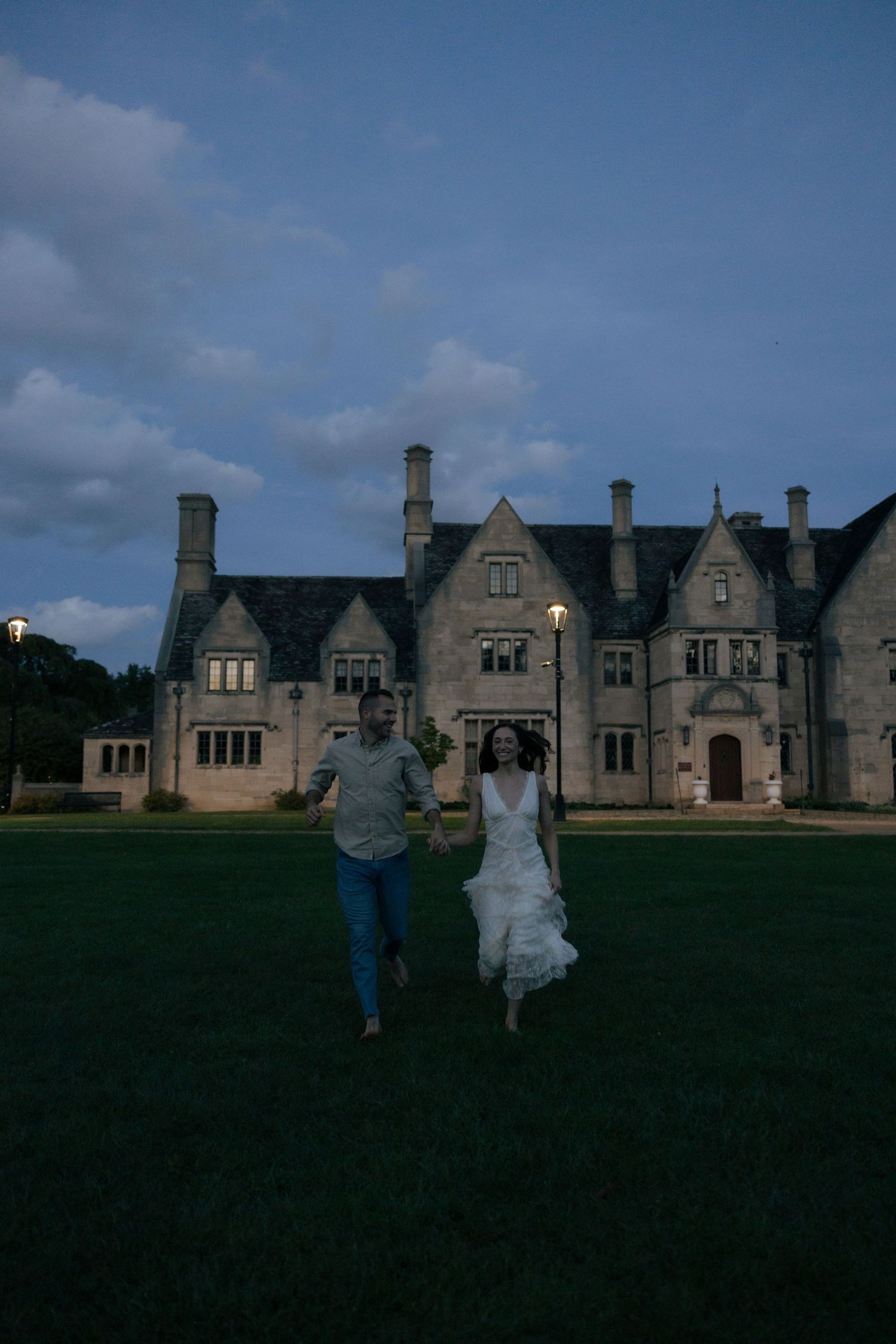 Joyful couple running hand in hand toward camera at dusk in front of a grand historic stone mansion.