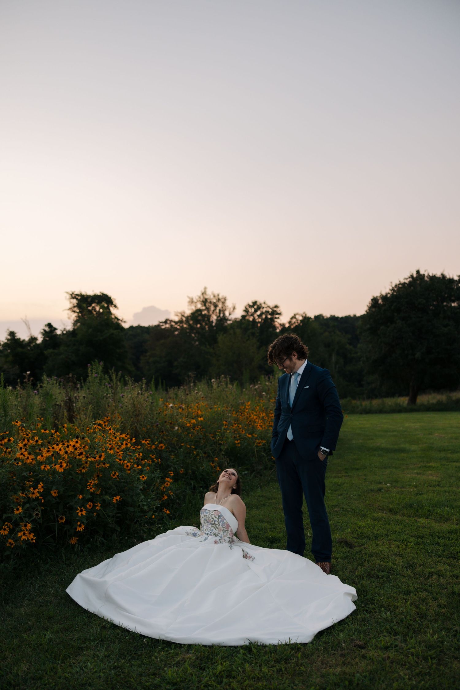 Bride in full white ballgown sits in grass as groom in navy suit looks down at her near wildflowers.