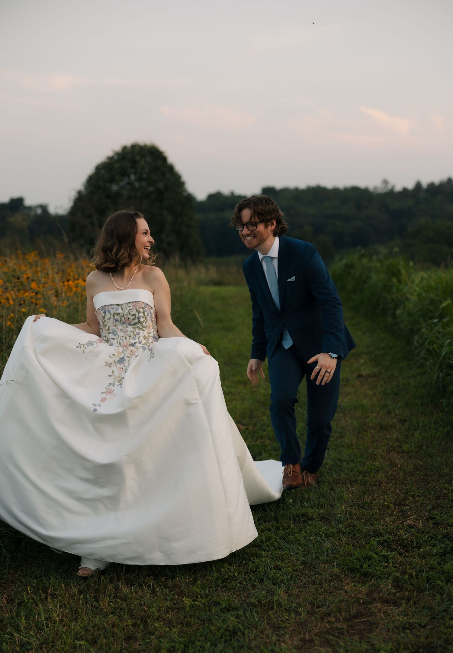 Laughing bride and groom stroll through lush green meadow at dusk, sharing a joyful wedding moment.