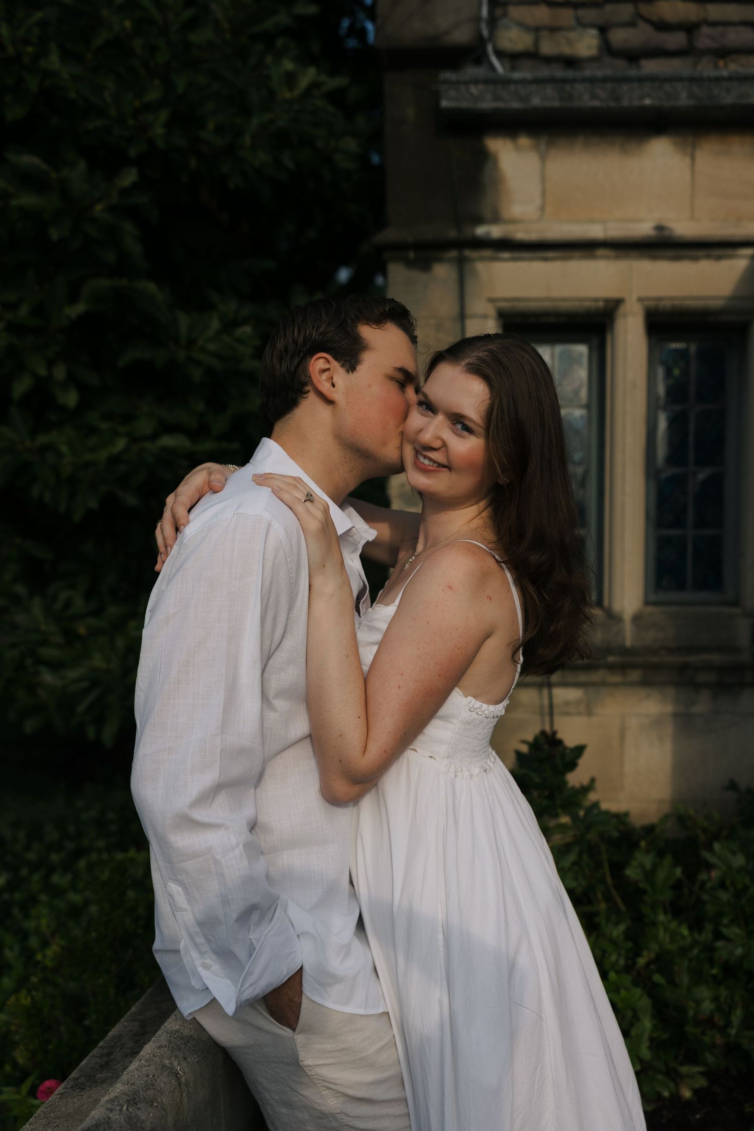 Couple in white embracing outdoors near stone building, man kissing woman's cheek as she smiles.