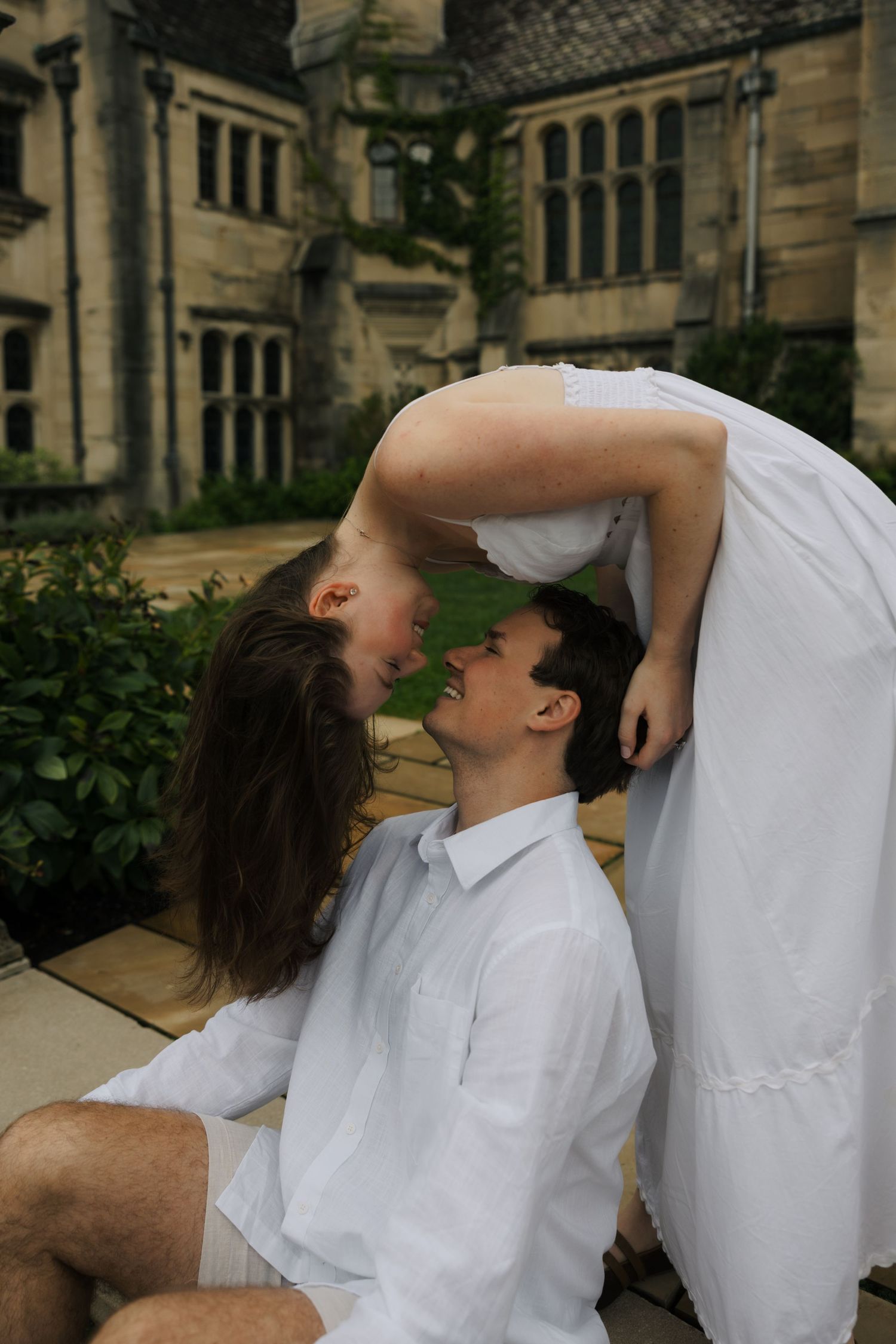 Romantic couple shares a kiss outside a historic stone manor, woman leaning over seated man.