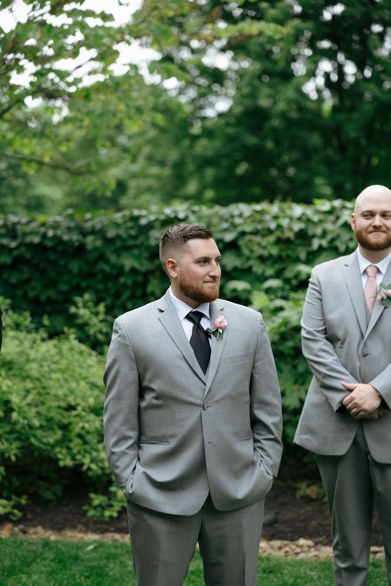 Groom in gray suit with pink boutonniere smiling during outdoor wedding ceremony surrounded by lush greenery.