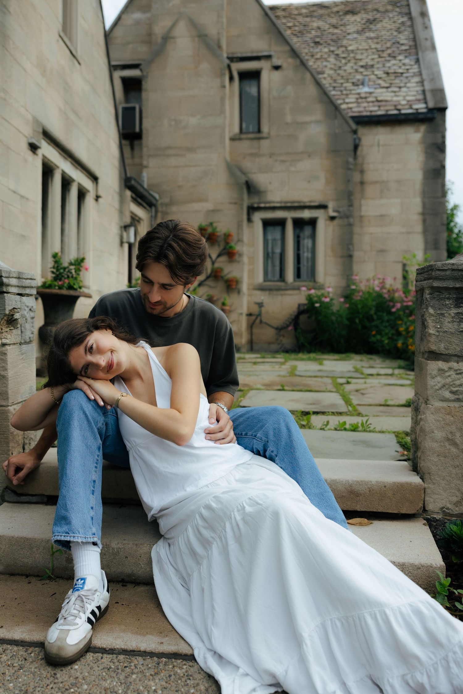 A couple sits on stone steps outside a historic building, the woman in a white dress resting her head on the man's shoulder.