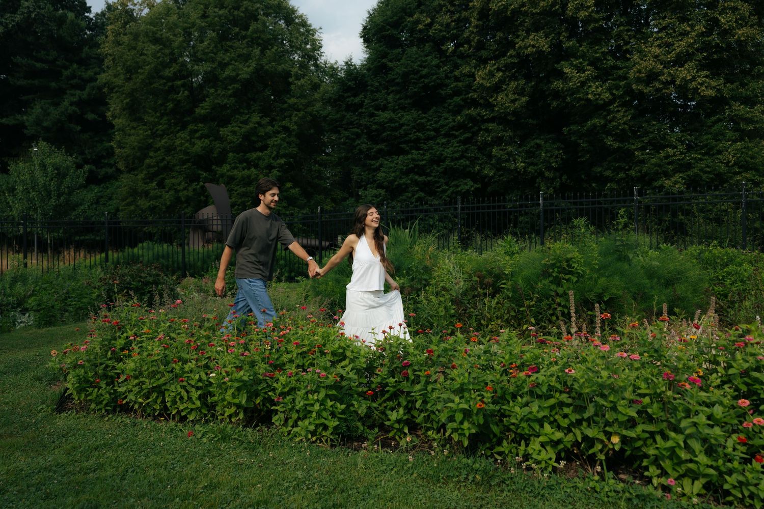 Couple holding hands walking through a lush garden with colorful flowers and green trees.