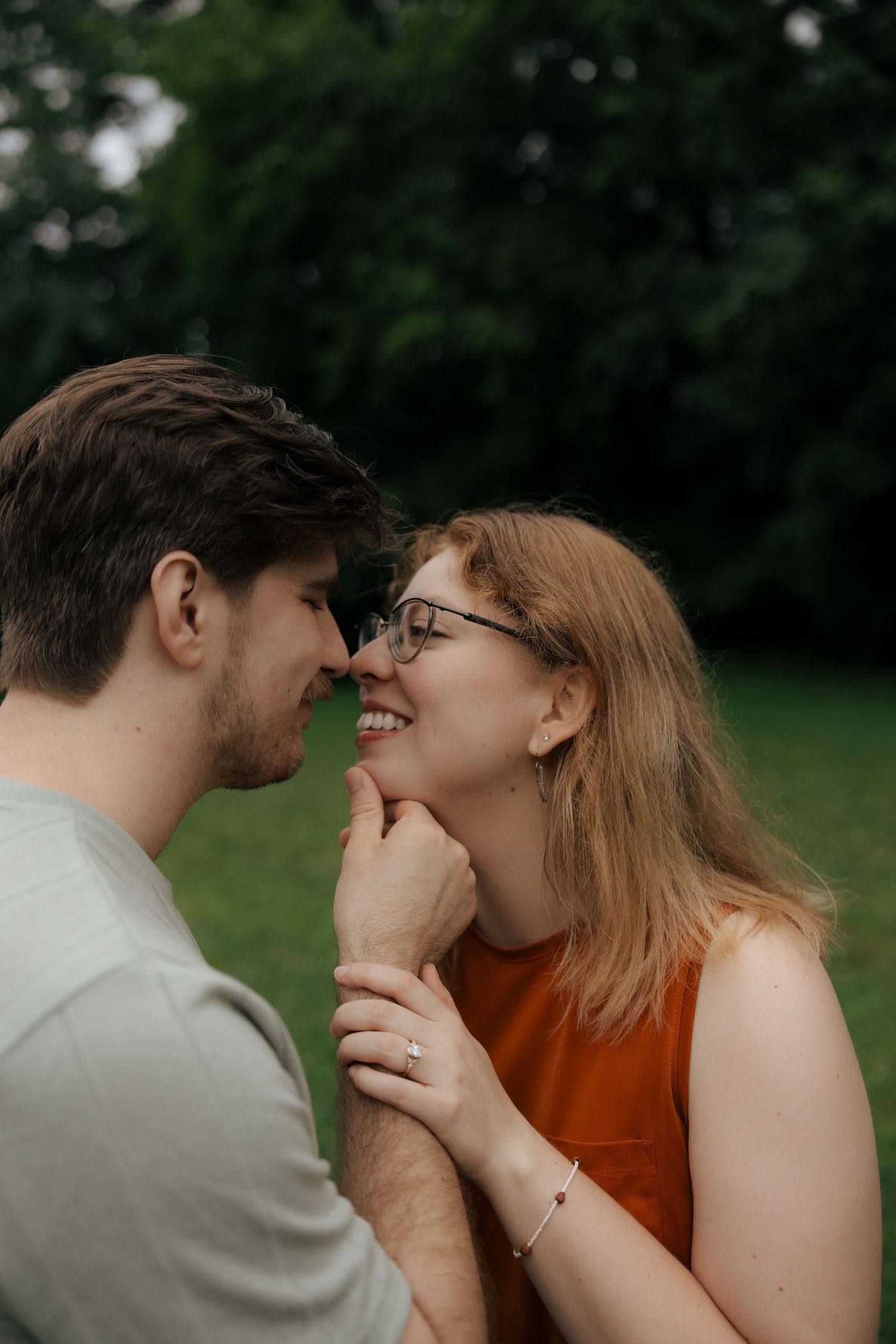 A smiling couple leans in close nose-to-nose in a tender moment outdoors in a green park.