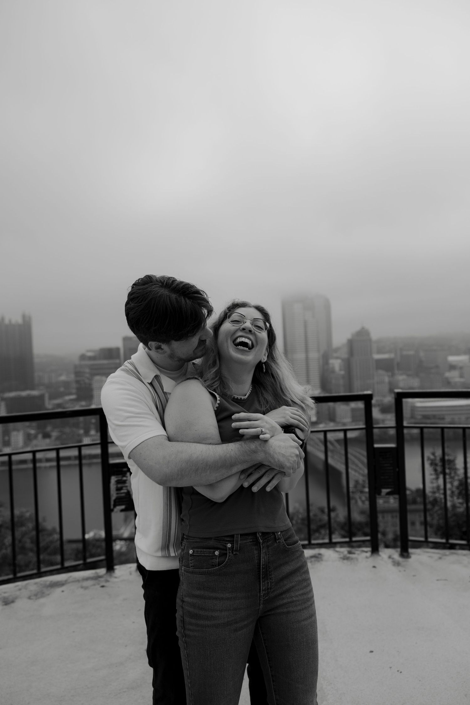 Black and white photo of a joyful couple embracing and laughing on a rooftop with city skyline behind them.
