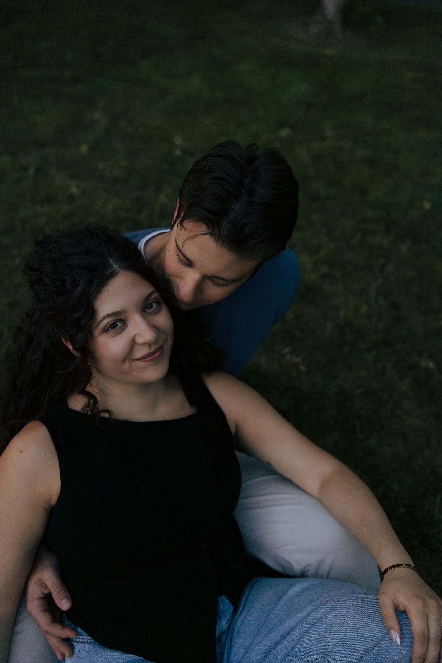Woman smiling as man tenderly rests his head against hers while sitting on grass.