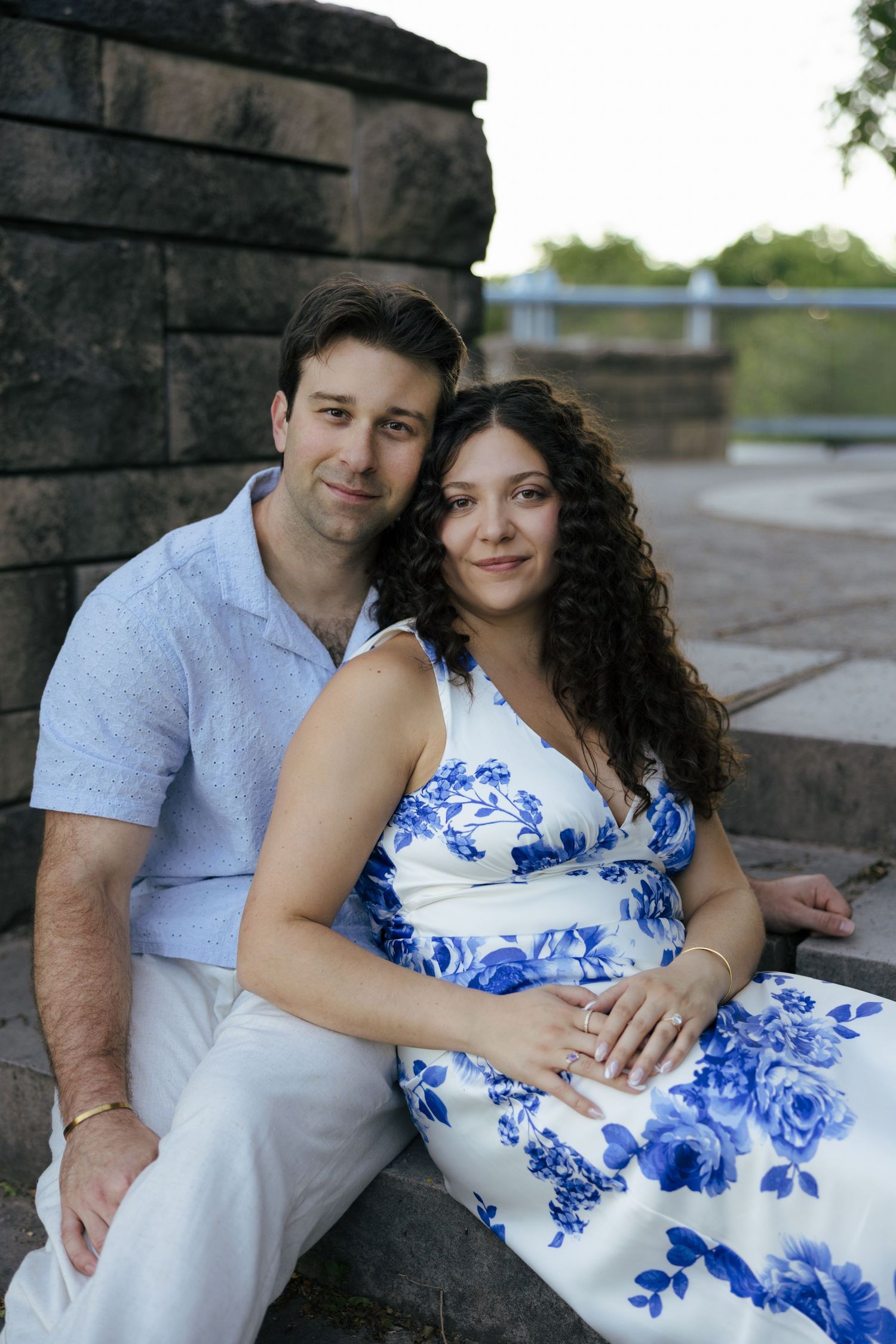 A couple sits together on stone steps, the woman in a blue floral dress leaning against her partner outdoors.
