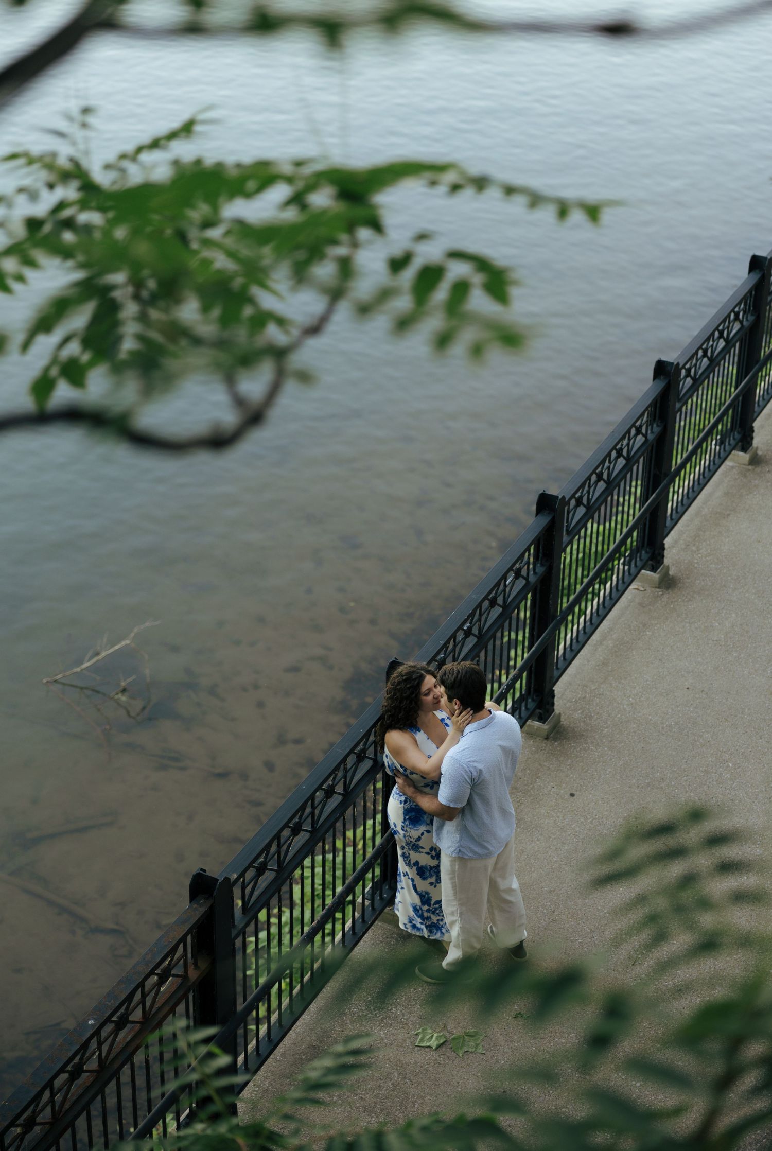 Aerial view of couple embracing along a riverside iron fence pathway framed by tree branches.