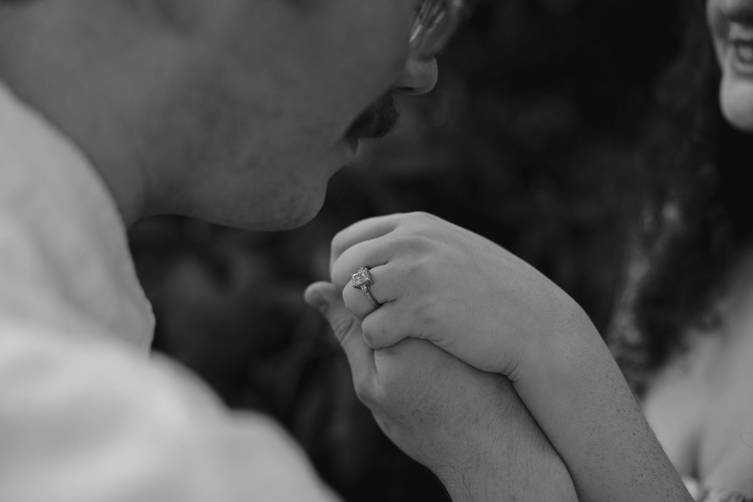 Black and white close-up of man kissing womans hand adorned with a sparkling engagement ring.