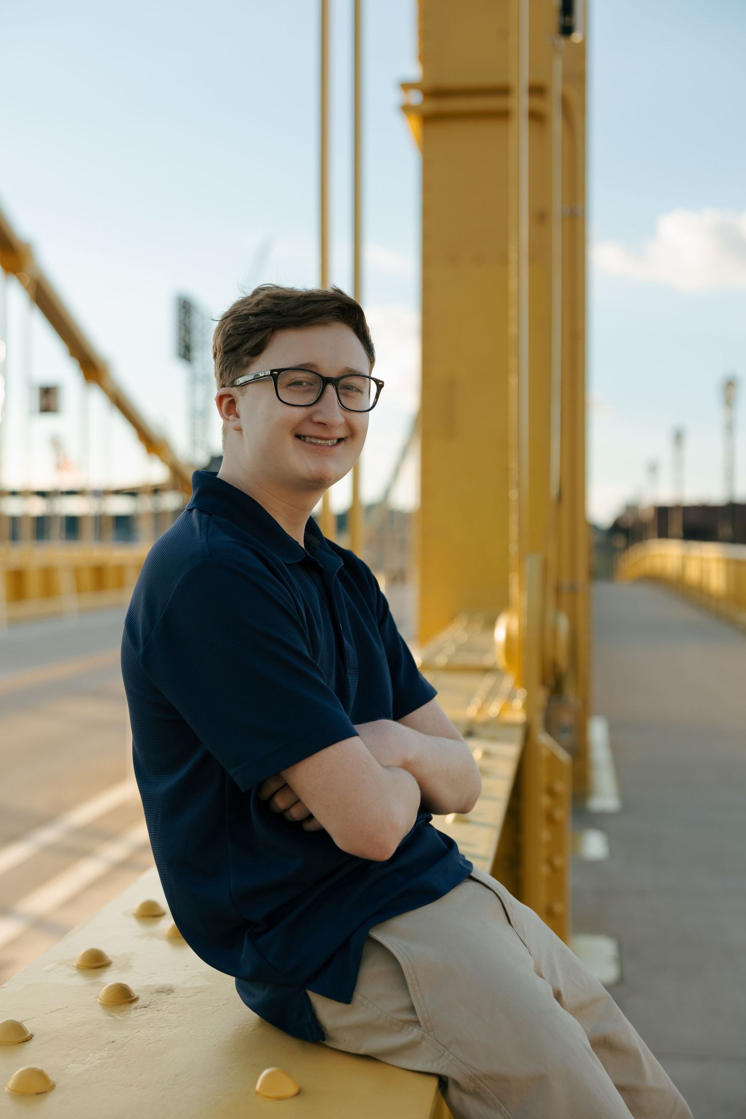 Teen boy in navy polo sitting on yellow bridge beam with arms crossed, smiling at camera outdoors.