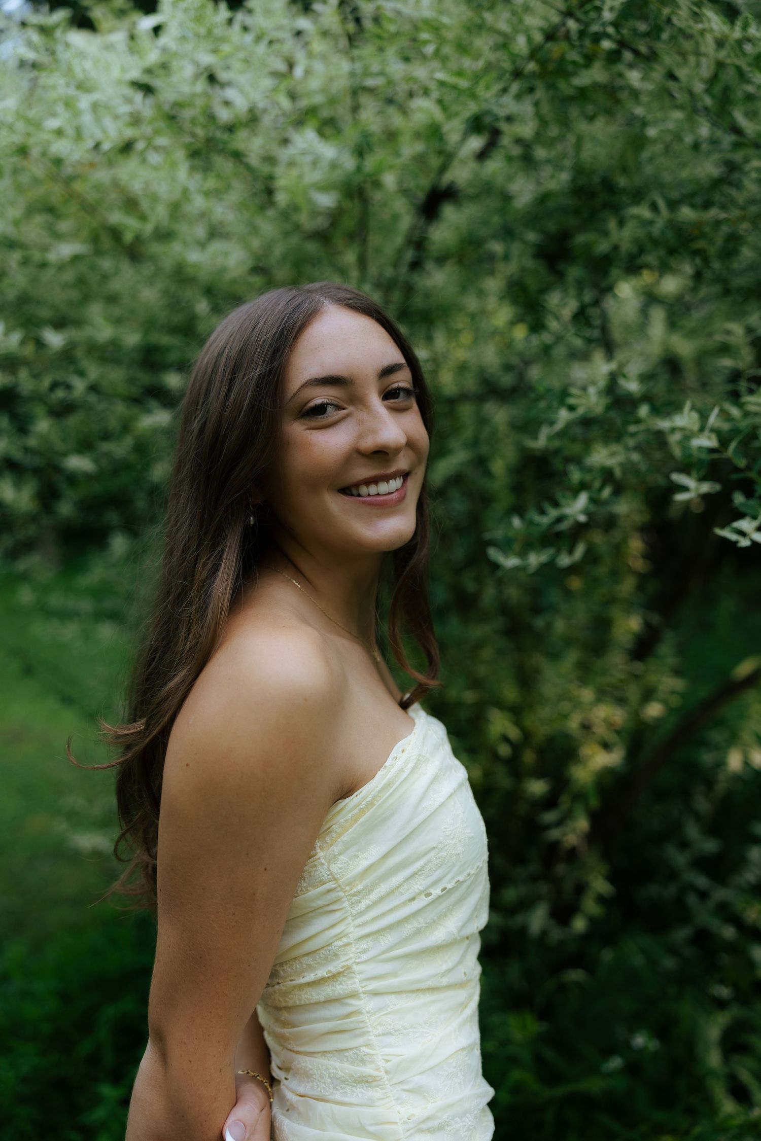 Smiling young woman in white strapless dress posing outdoors surrounded by green foliage and trees.