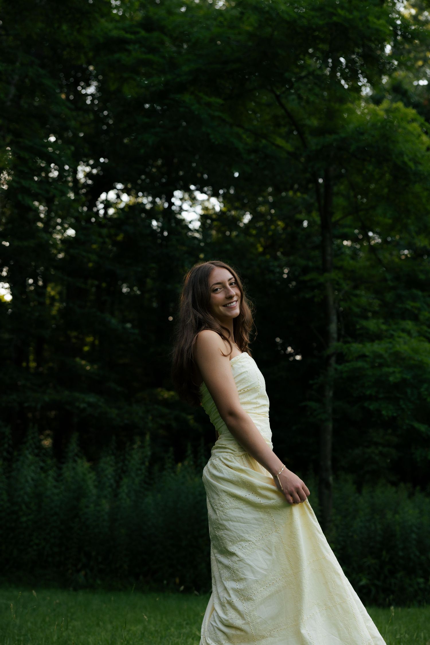Young woman in flowing yellow dress smiling outdoors in lush green park setting at dusk.