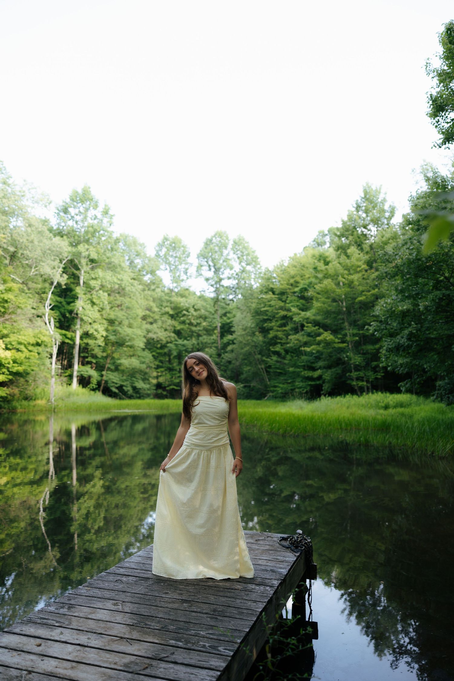 Young woman in flowing white dress standing on a wooden dock surrounded by a peaceful forest lake.