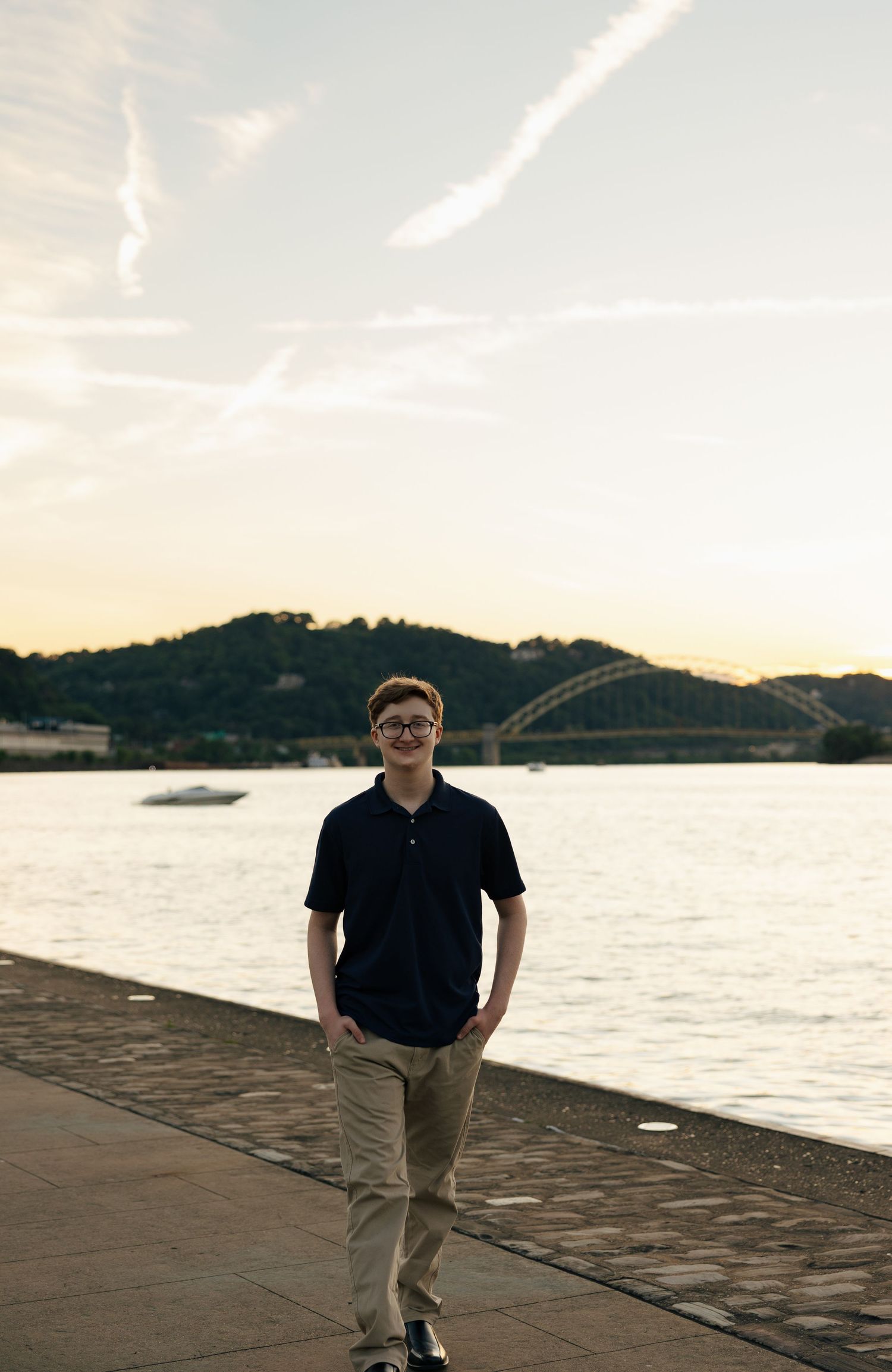 Young man in navy polo standing on riverside walkway with bridge and hills in background at sunset.