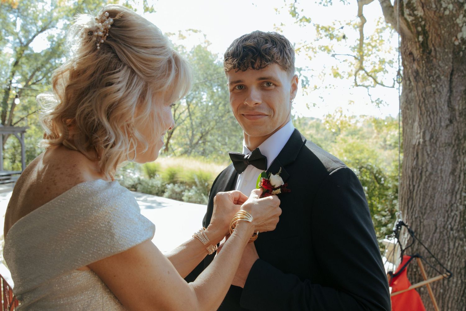 Mother pins boutonniere on groom's black tuxedo lapel outdoors before wedding ceremony.