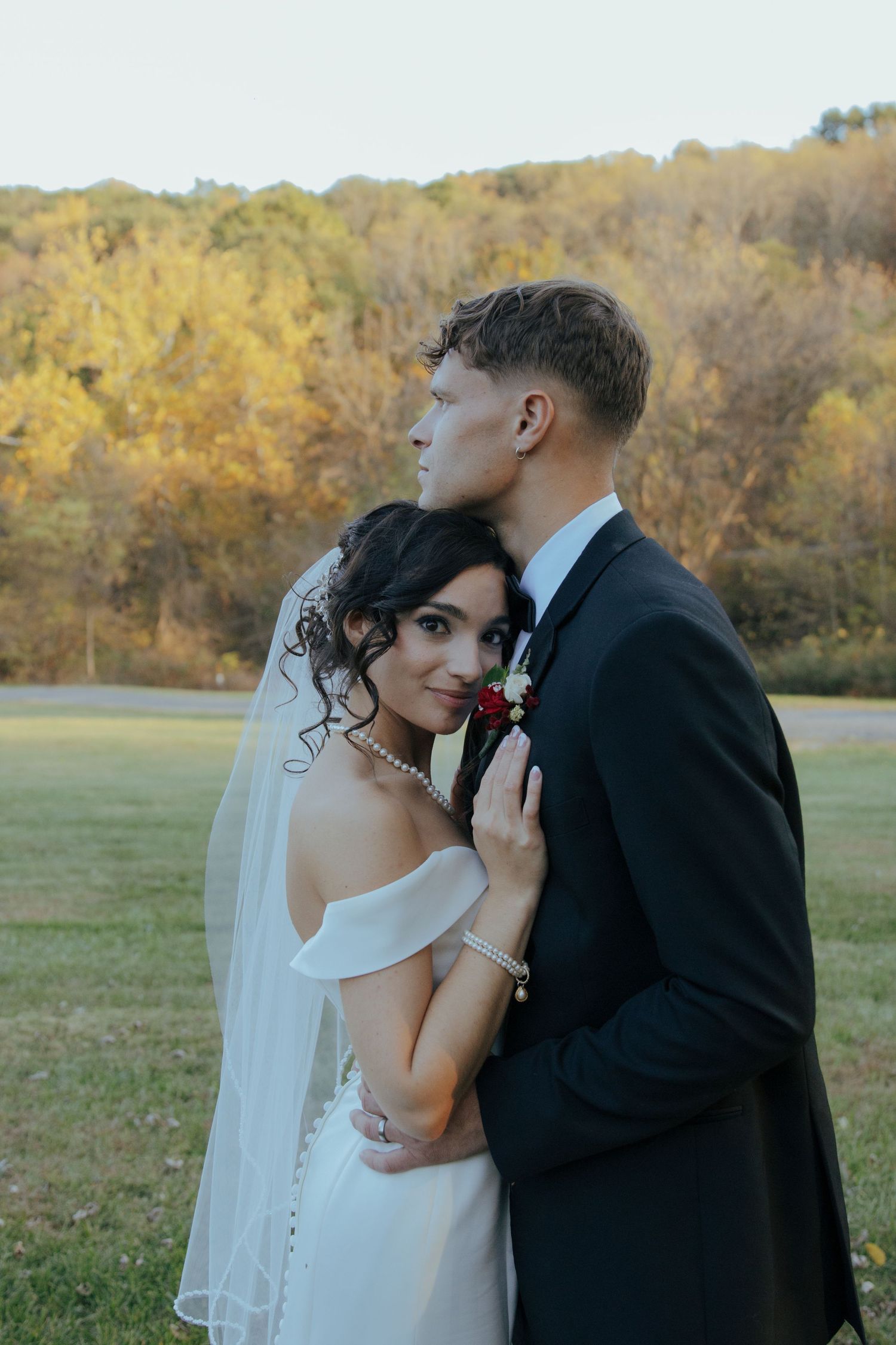 Groom in black suit kisses bride wearing white off-shoulder gown and veil outdoors in autumn setting.