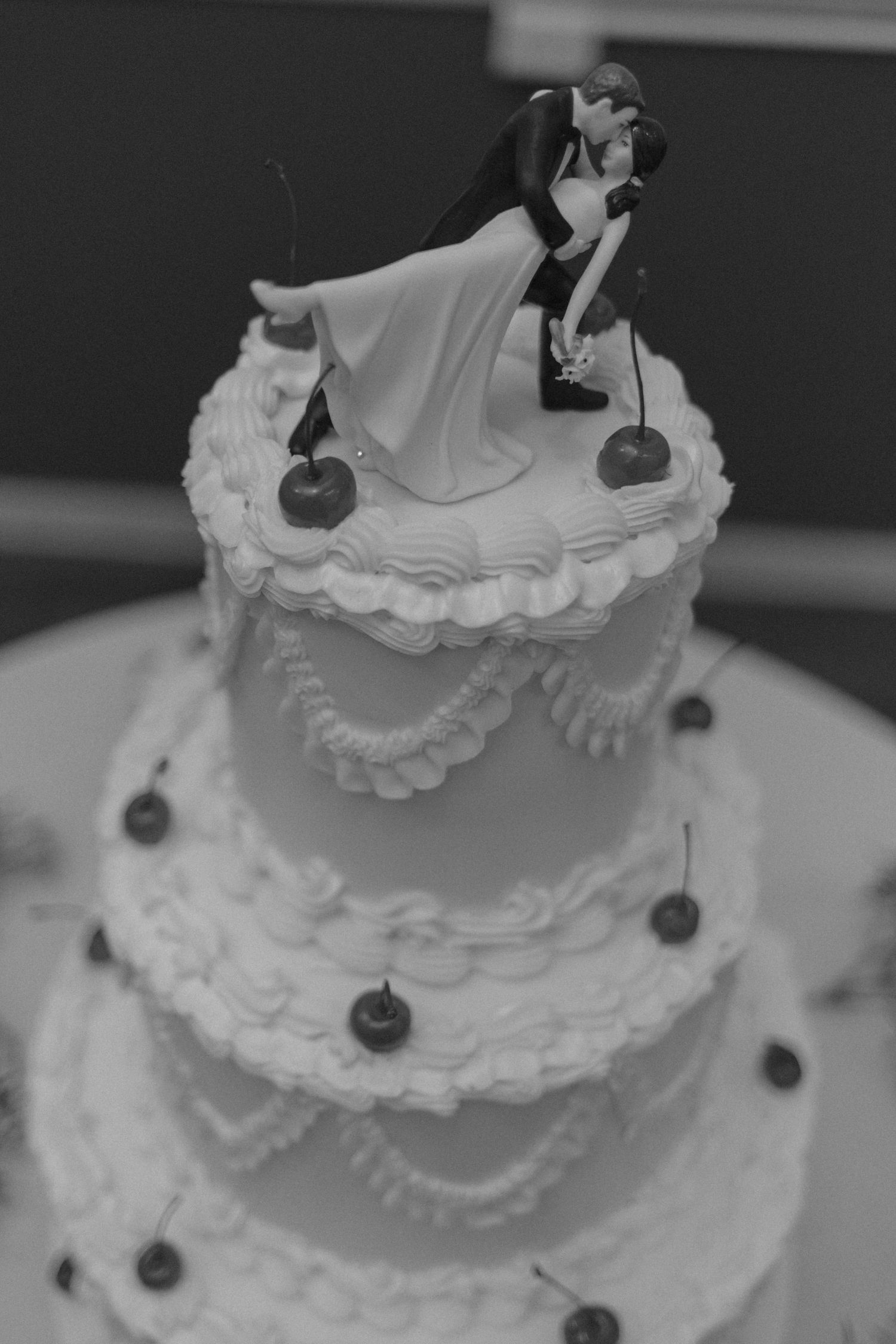Elegant three-tier white wedding cake with bride and groom topper and cherry decorations.