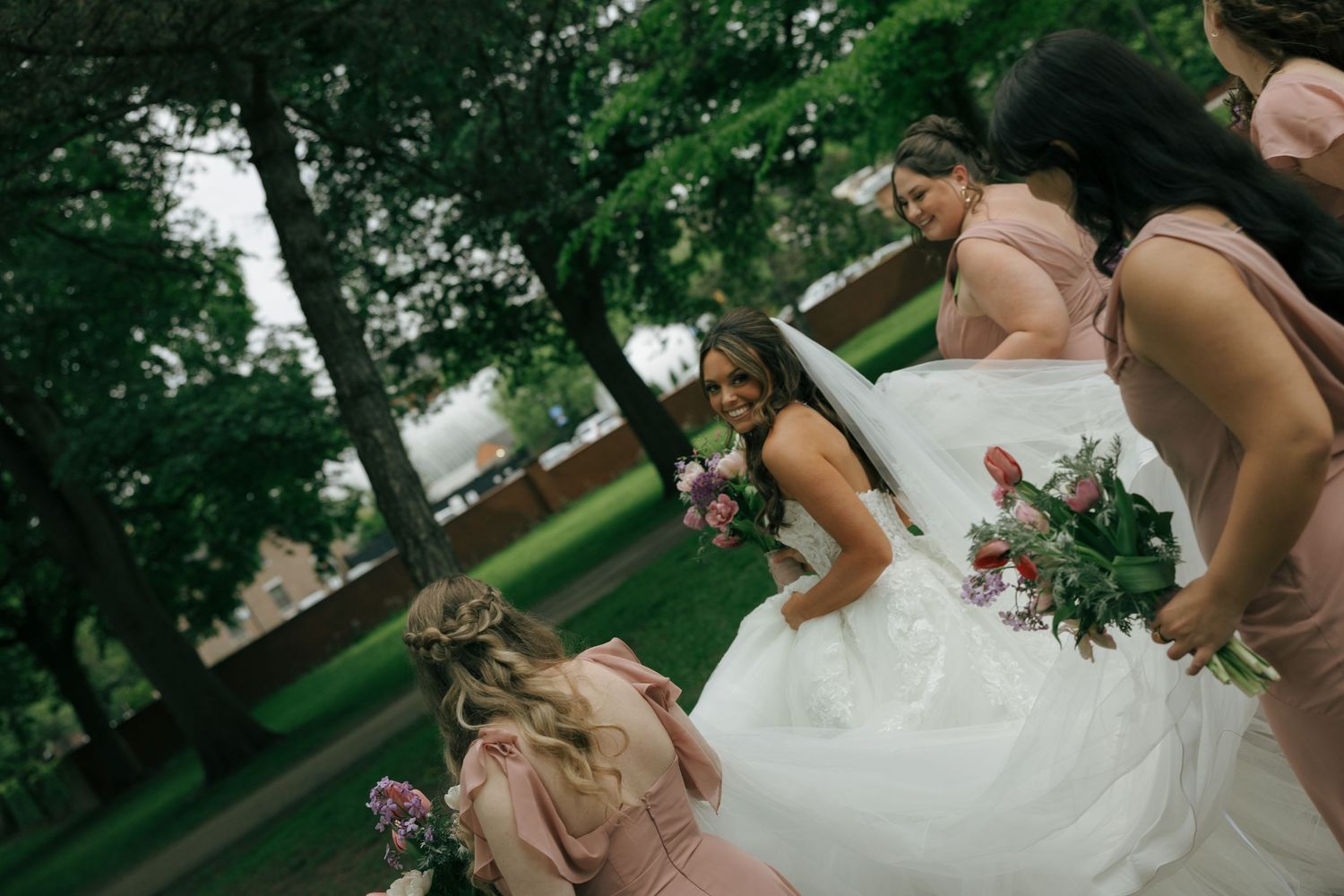 Bride in white ball gown smiles holding pink bouquet surrounded by bridesmaids in mauve dresses.