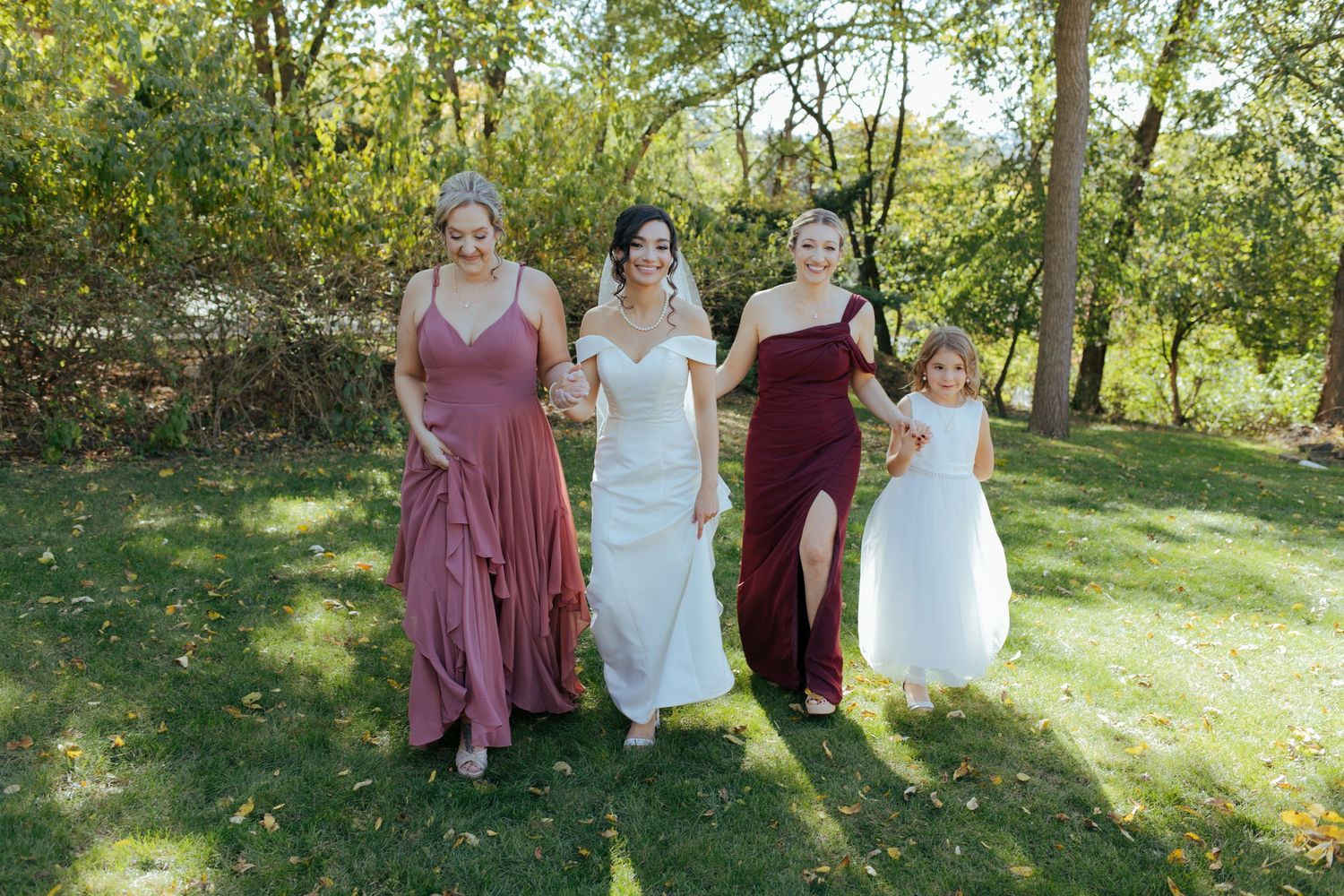 Bride in white gown walks with bridesmaids in burgundy and mauve dresses and flower girl in white outdoors.