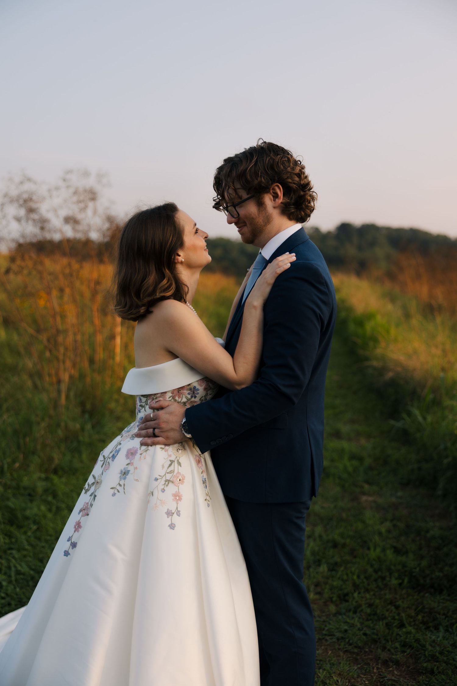 Bride and groom embrace at golden hour in a field, she wears a floral embroidered off-shoulder ball gown.