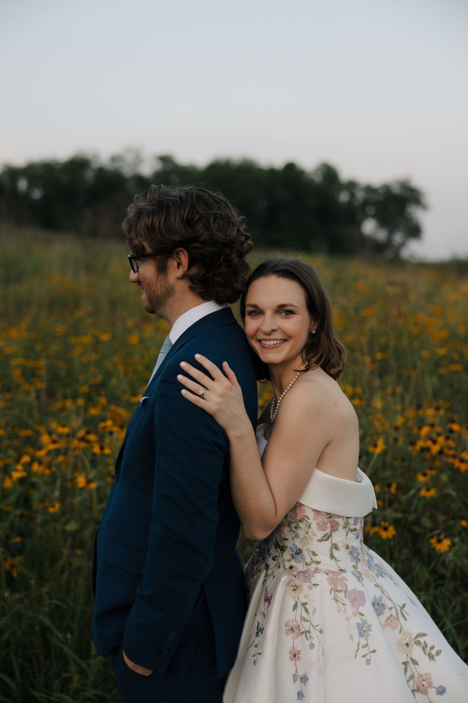 Smiling bride in a floral wedding gown embracing groom from behind in a field of yellow wildflowers at sunset.